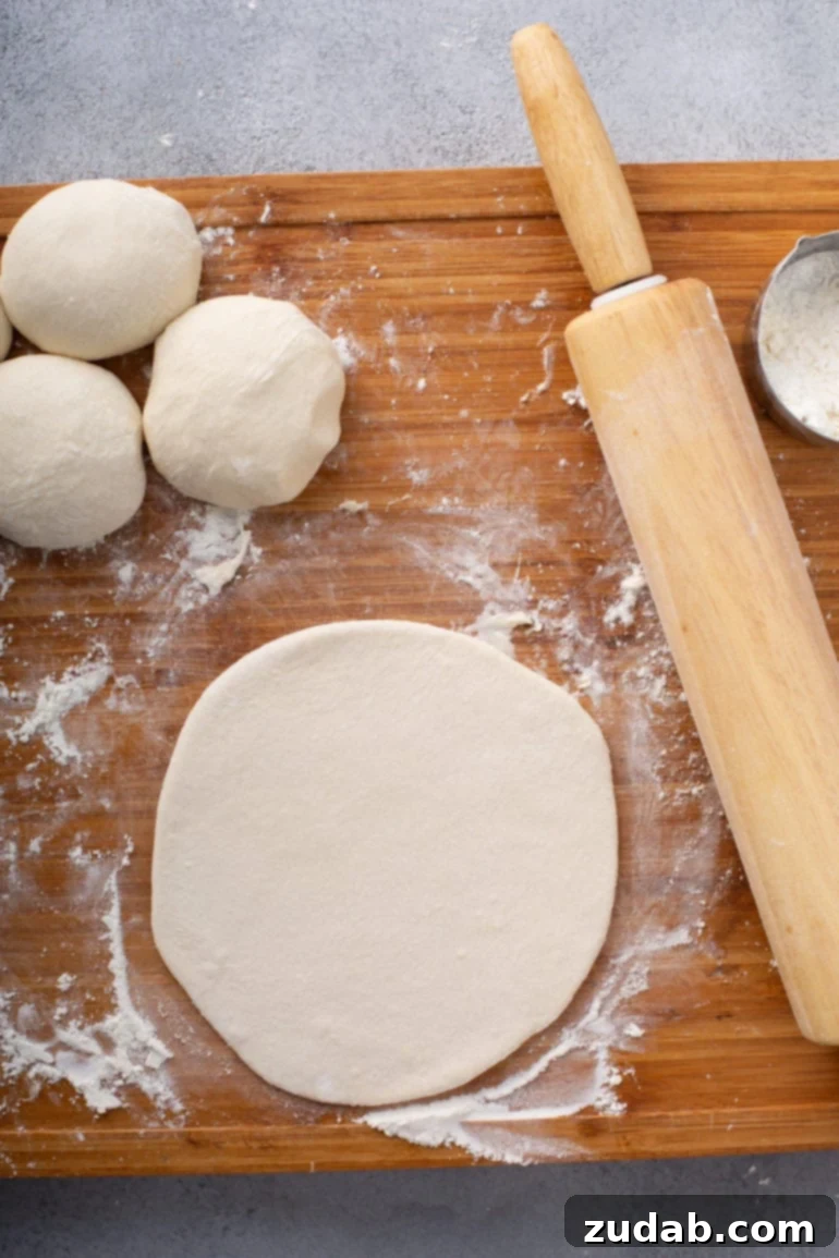 Whip Up Three Ingredient Unleavened Bread 4 A photo of a portion of dough rolled out into a flat circle on a floured wooden cutting board with a wooden rolling pin sitting next to it and unrolled portions of dough also sitting next to it.