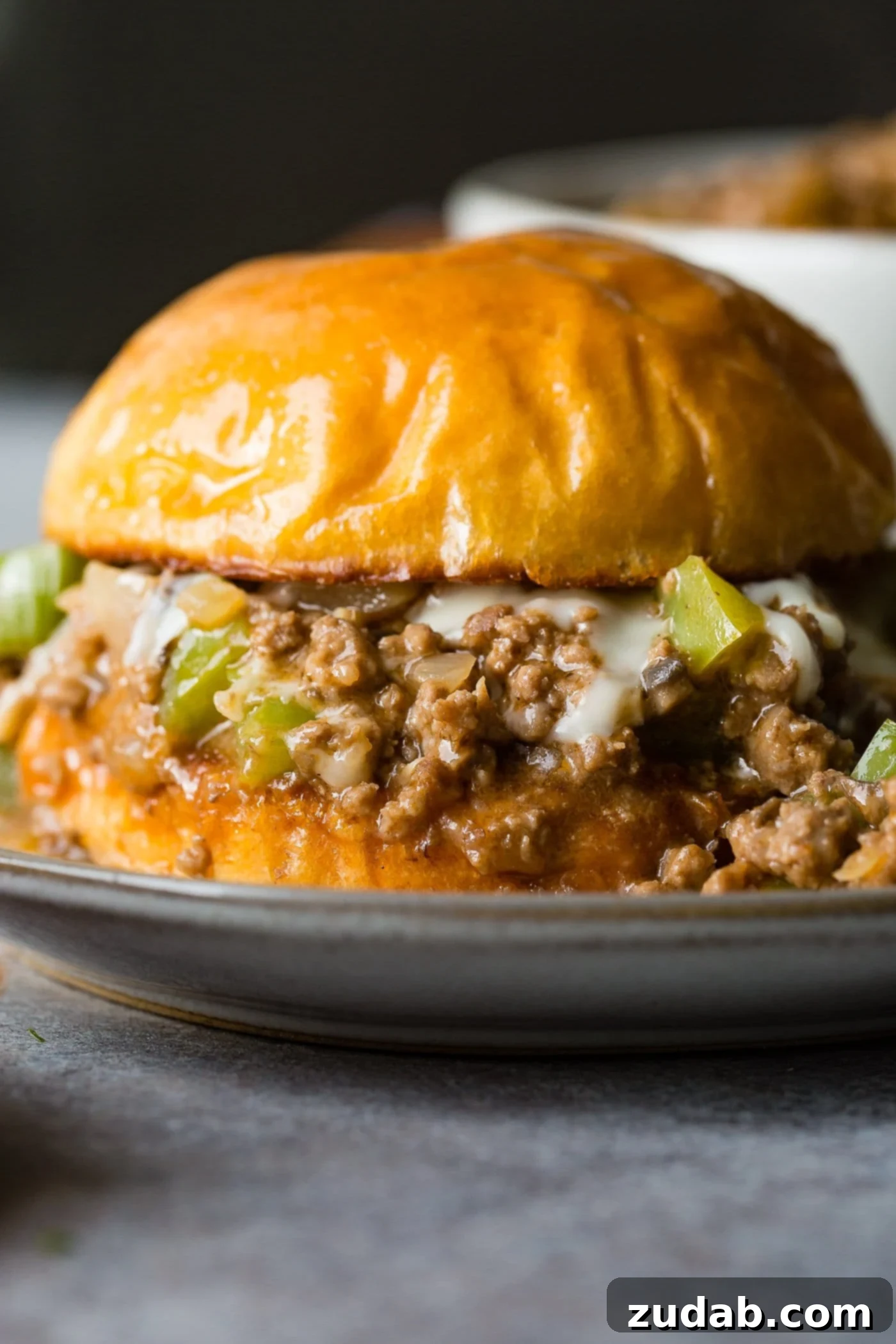 Philly Cheesesteak Sloppy Joes A photo of a philly cheesesteak sloppy joe on a toasted brioche bun on a white plate.