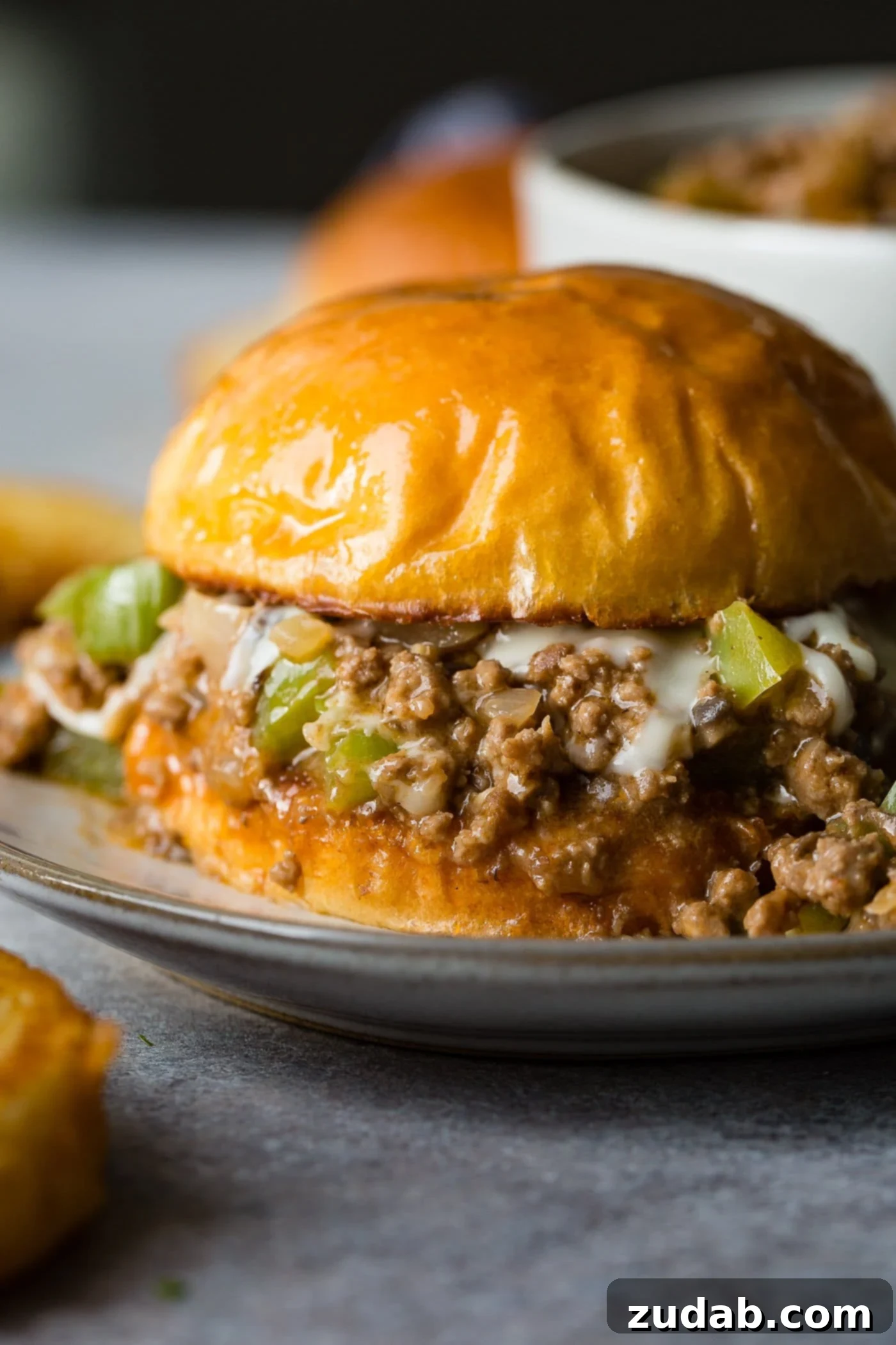 Philly Cheesesteak Sloppy Joes A photo of a philly cheesesteak sloppy joe on a toasted brioche bun on a white plate.