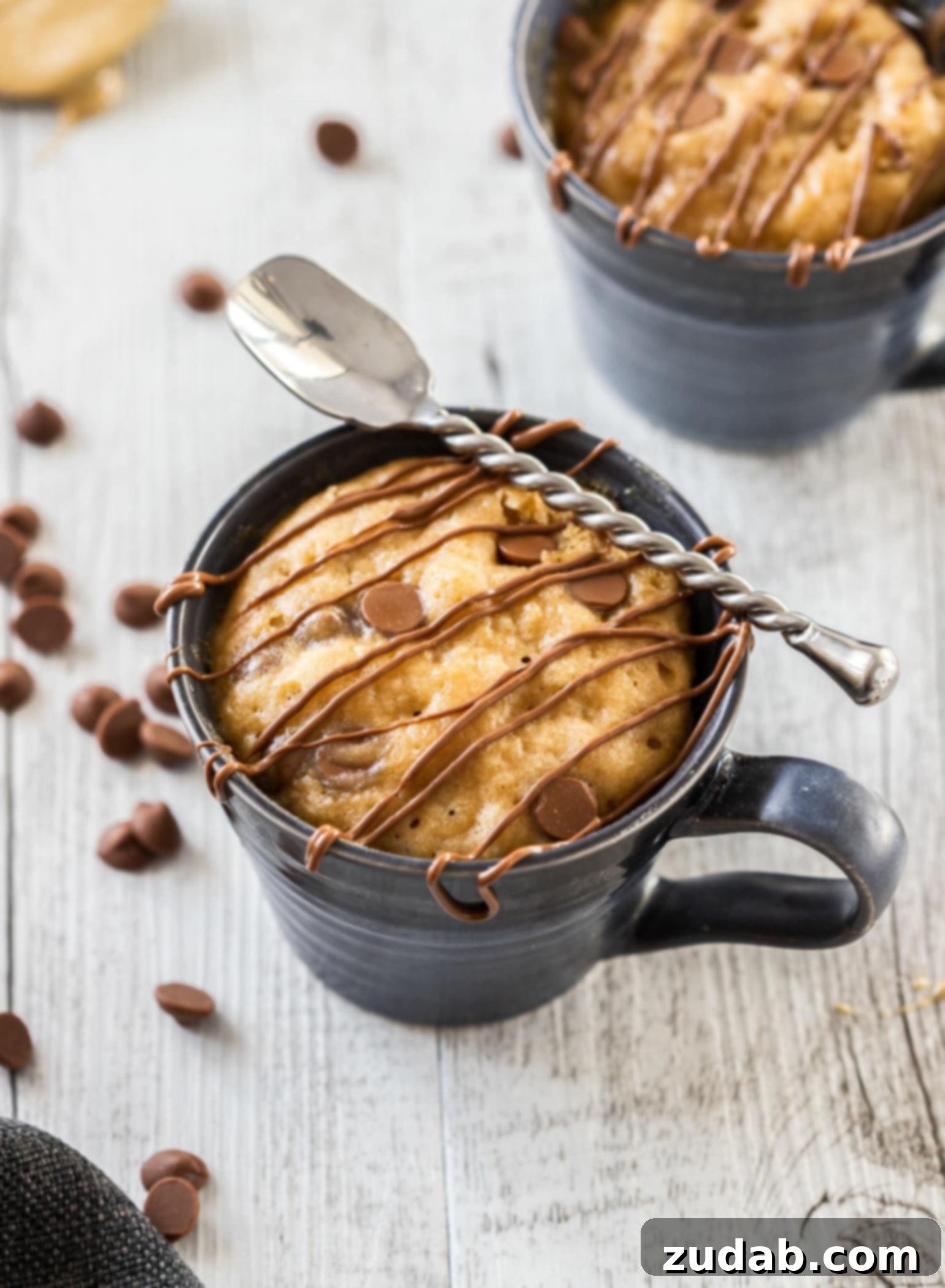 Microwave PB Bliss 2 Birdseye view of a mug cake in a blue mug with chocolate drizzled over the top and a spoon sitting on top, emphasizing its deliciousness and ease of preparation.