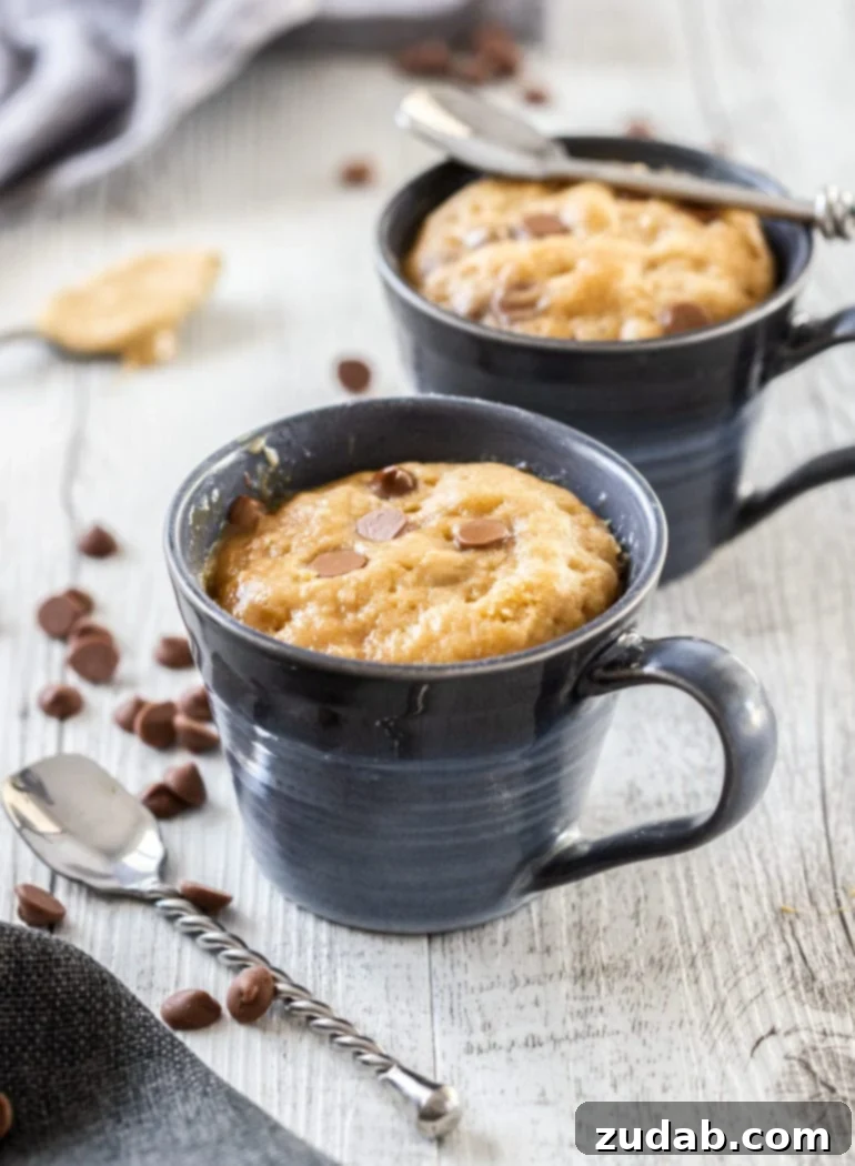 Microwave PB Bliss 3 Two blue mugs filled with cake and surrounded by chocolate chips and spoons, highlighting the ease and individual serving size of mug cakes.