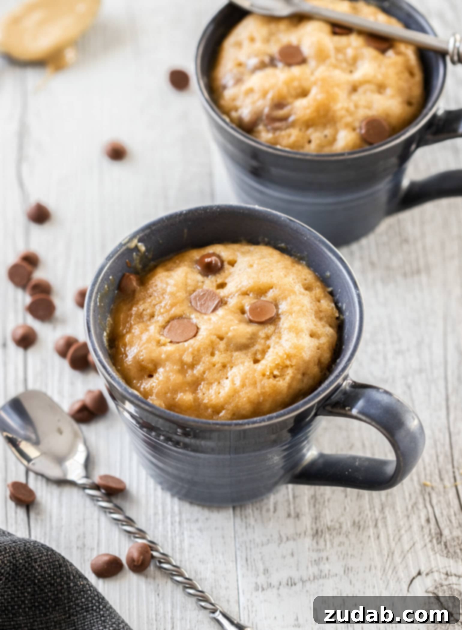 Microwave PB Bliss 6 Two blue mugs filled with cake and surrounded by chocolate chips and spoons, highlighting the ease and deliciousness of microwave mug cakes for late-night cravings.