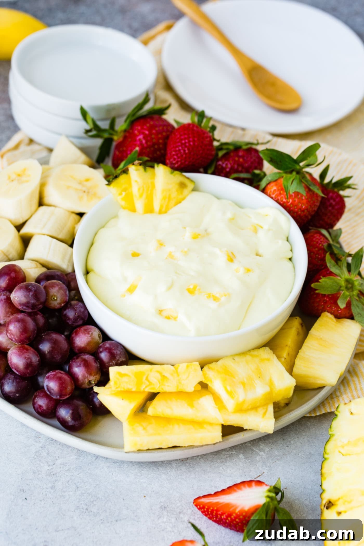Luscious Pineapple Cream Cheese Fruit Dip 2 A bowl of pineapple cream cheese fruit dip with three pieces of pineapple on the edge of the bowl. The bowl is sitting on a serving tray with banana slices, red grapes, pineapple chunks, and strawberries surrounding the bowl.