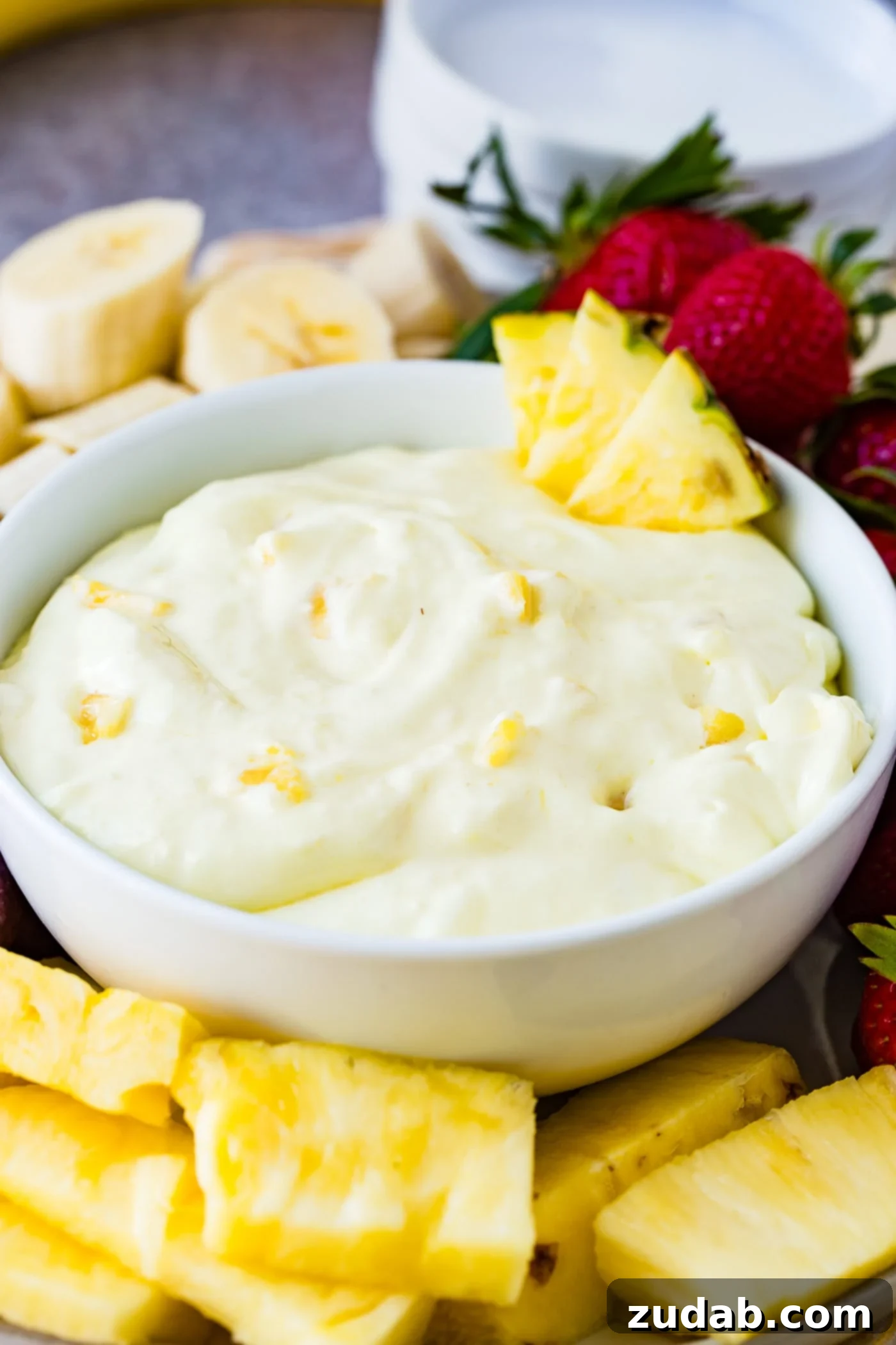 Luscious Pineapple Cream Cheese Fruit Dip 3 A white bowl with pineapple cream cheese fruit dip. There are four pineapple chunks on the edge of the bowl and little bits of pineapple in the dip. The bowl is surrounded with sliced bananas, whole strawberries, grapes, and pineapple chunks.