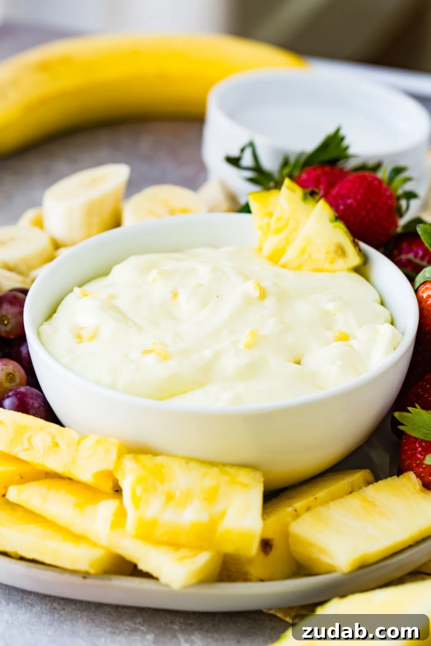 Luscious Pineapple Cream Cheese Fruit Dip 4 A white bowl containing pineapple fruit dip. There are three chunks of pineapple on the edge of the bowl and tiny pieces of pineapple in the white dip. Sliced bananas, pineapple chunks, and wholes strawberries and red grapes surround the bowl. A banana and a stack of dessert plates are in the background.