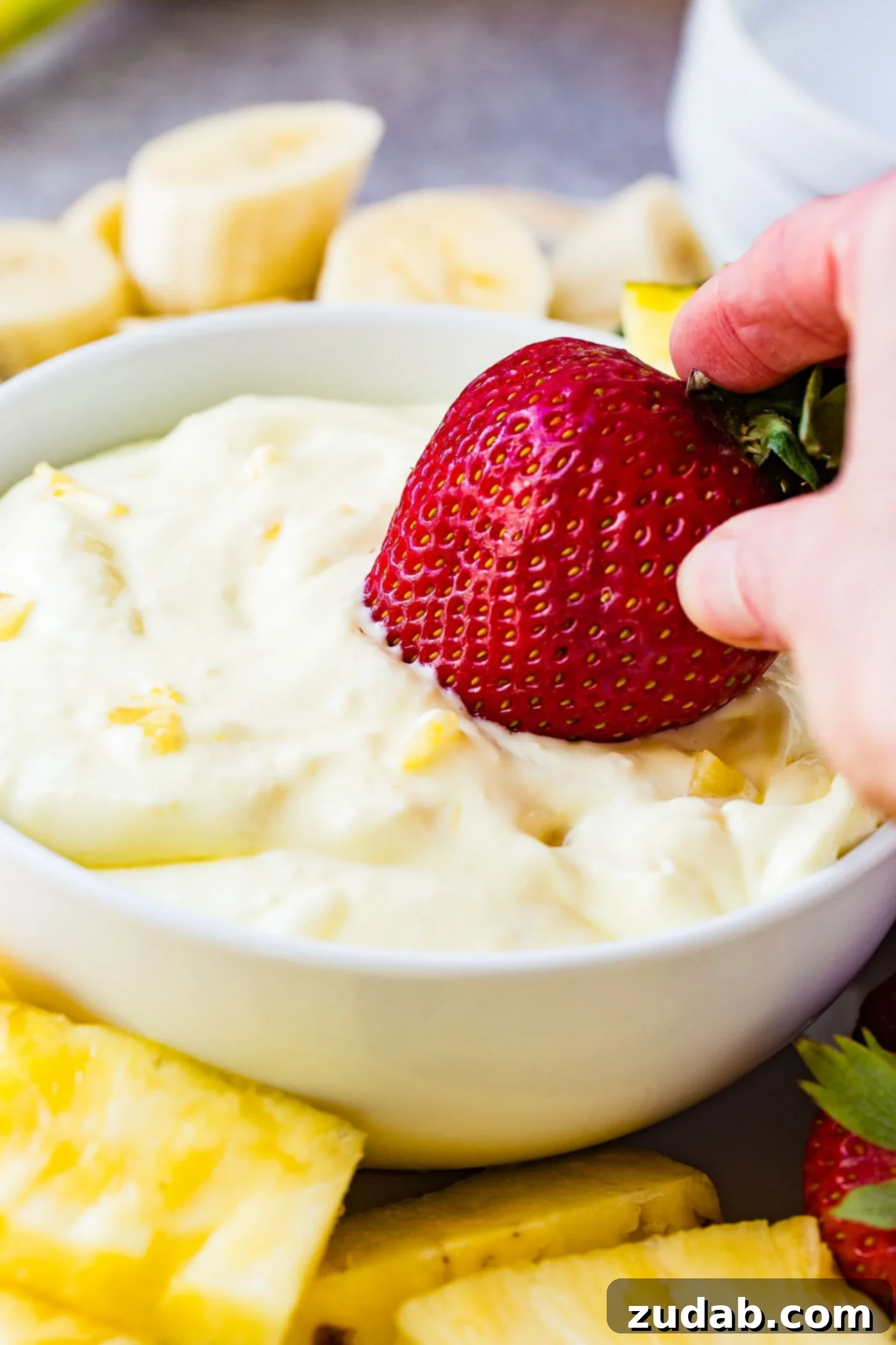 Luscious Pineapple Cream Cheese Fruit Dip 5 A whole red strawberry being dipped in a bowl of pineapple fruit dip with tiny pieces of pineapple in the dip. There are sliced bananas, and pineapple chunks surrounding the bowl of dip.