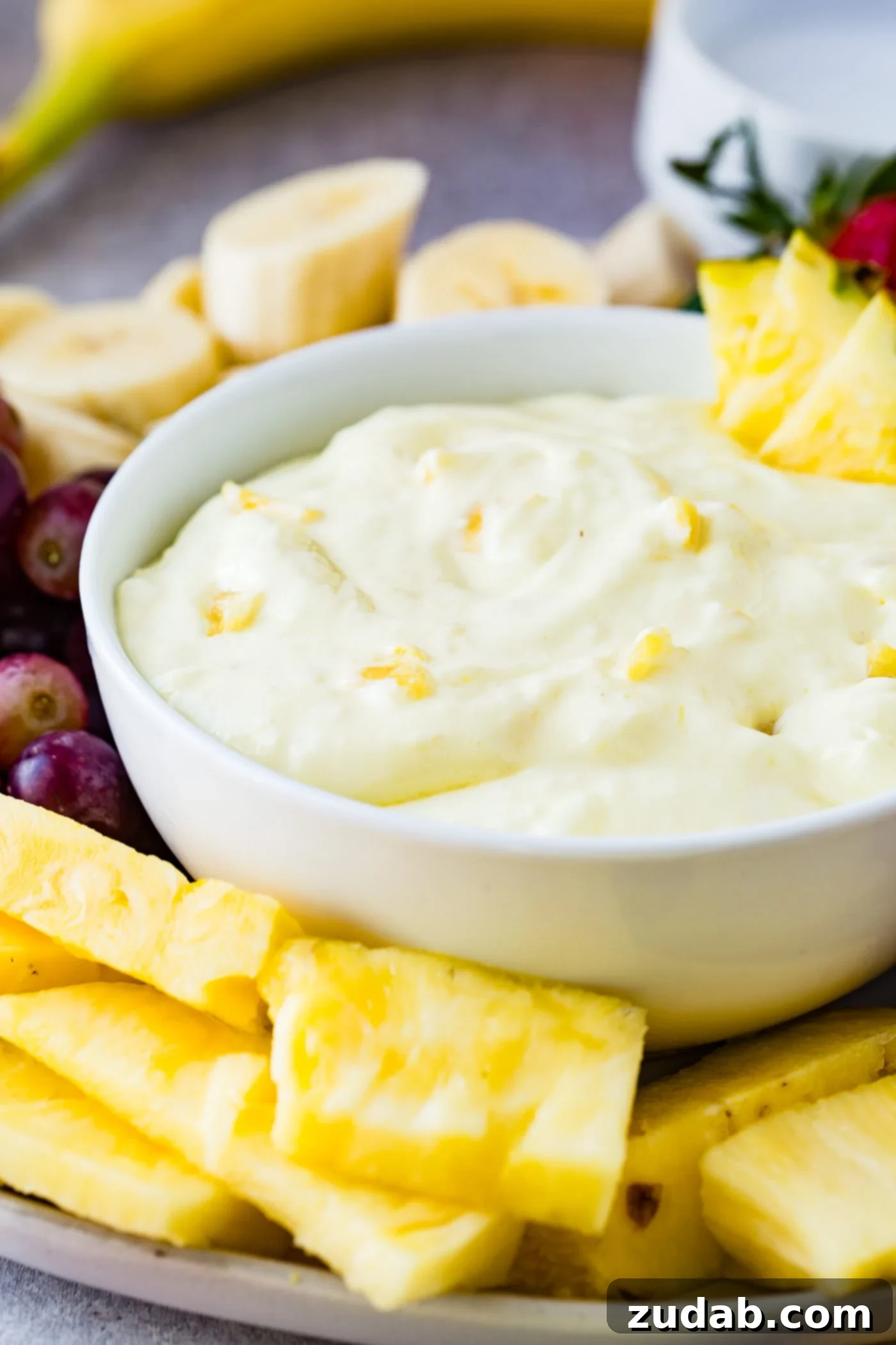 Luscious Pineapple Cream Cheese Fruit Dip 6 A bowl of pineapple fruit dip with three pineapple chunks on the edge of the bowl and tiny pieces of pineapple in the dip. The bowl is surrounded by sliced bananas, red grapes, pineapple chunks and whole strawberries.