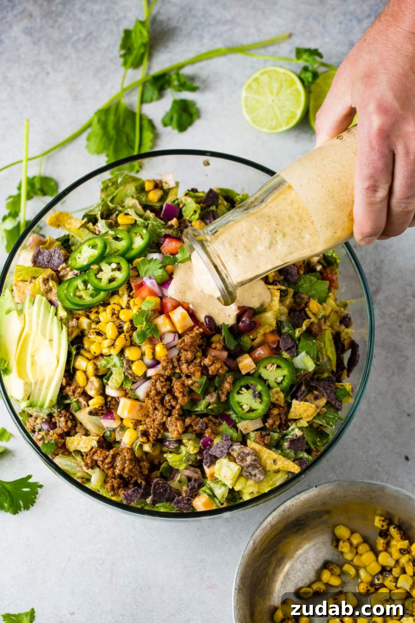 A glass bowl generously filled with Tex-Mex taco salad, featuring inviting avocado slices, golden corn, savory taco meat, tangy tomatillos, juicy tomatoes, and crunchy chips, as a rich dressing is poured from a glass container over the vibrant ingredients.