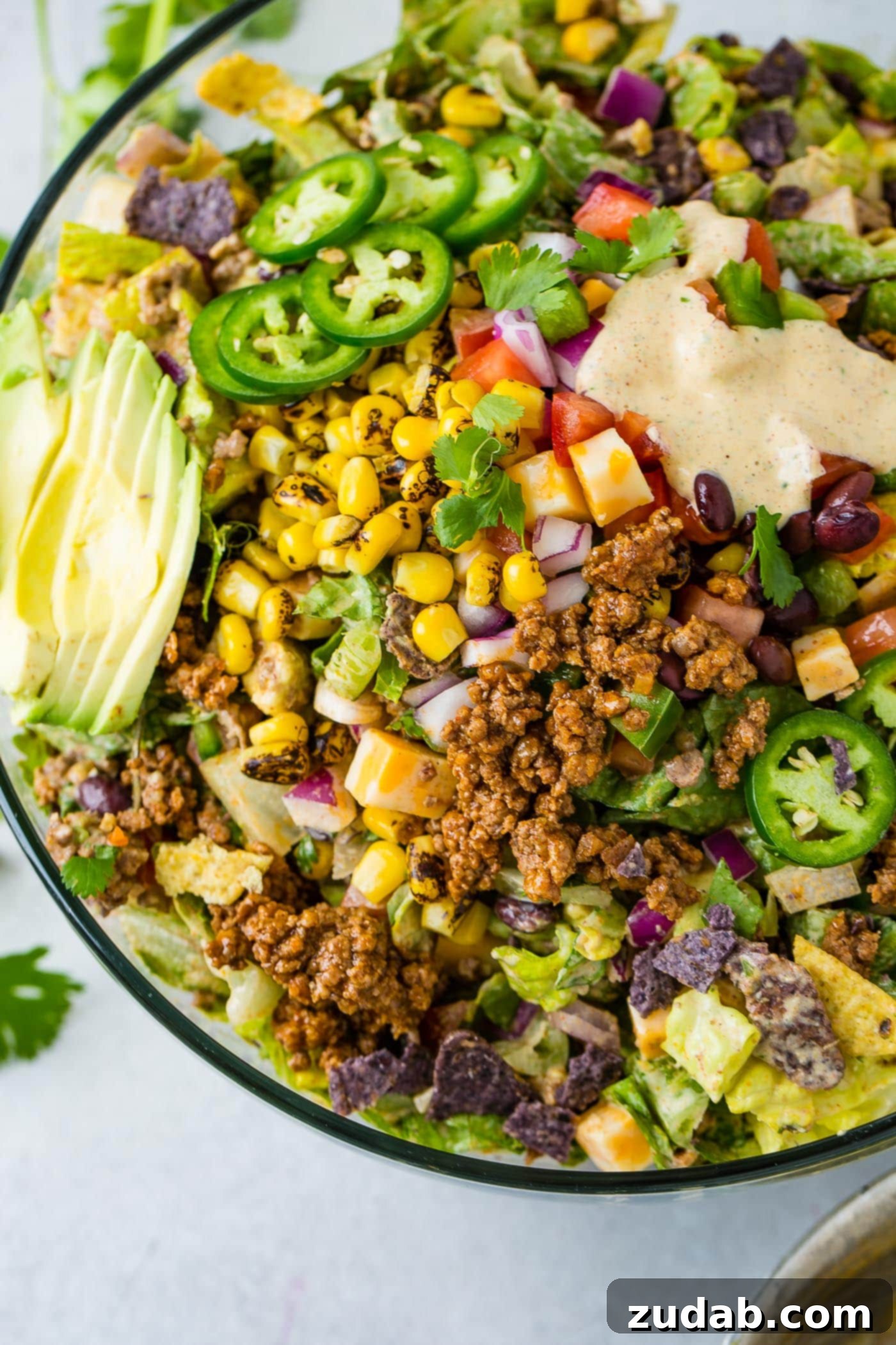 A large, inviting bowl of Tex-Mex taco salad, brimming with colorful ingredients including tangy tomatillos, crisp lettuce, sweet roasted corn, fresh cilantro, seasoned ground beef, and crunchy tortilla chips, with a subtle drizzle of dressing in the corner.