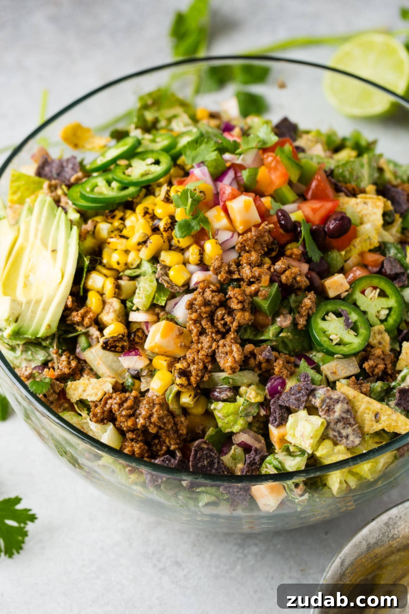 A vibrant glass bowl of Tex-Mex taco salad featuring crisp lettuce, seasoned ground beef, roasted corn, fresh tomatillos and tomatoes, topped with creamy sliced avocado.
