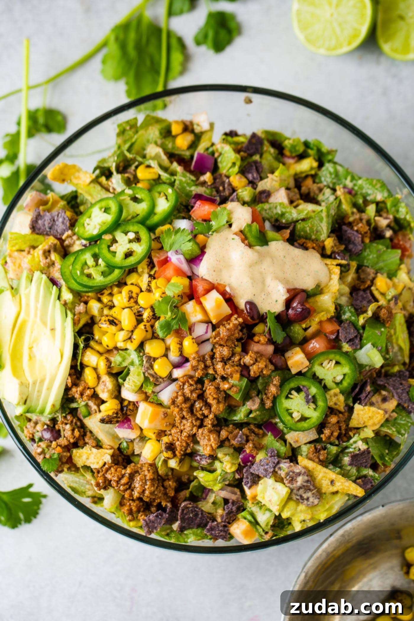 A top-down view of a large glass bowl showcasing a colorful Tex-Mex taco salad, richly layered with crisp lettuce, golden corn, creamy avocado slices, tangy tomatillos, seasoned taco meat, and a drizzle of dressing.