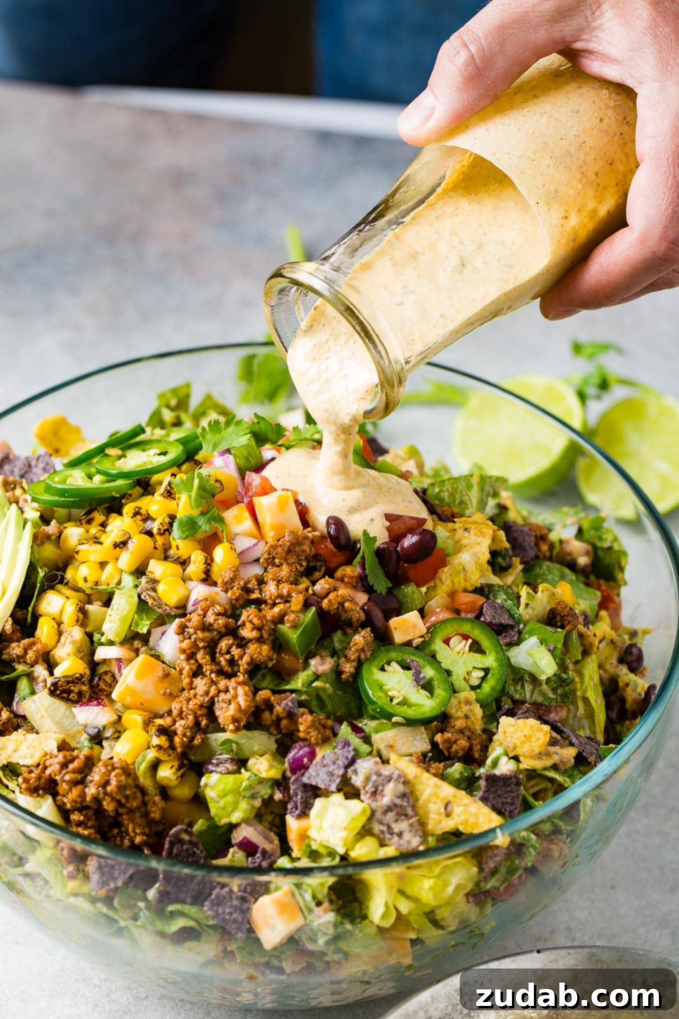 A glass bowl filled with vibrant Tex-Mex taco salad, with a rich, creamy dressing being poured over the top. The salad is generously packed with roasted corn, tangy tomatillos, juicy tomatoes, crisp lettuce, seasoned taco meat, and elegantly sliced avocado.