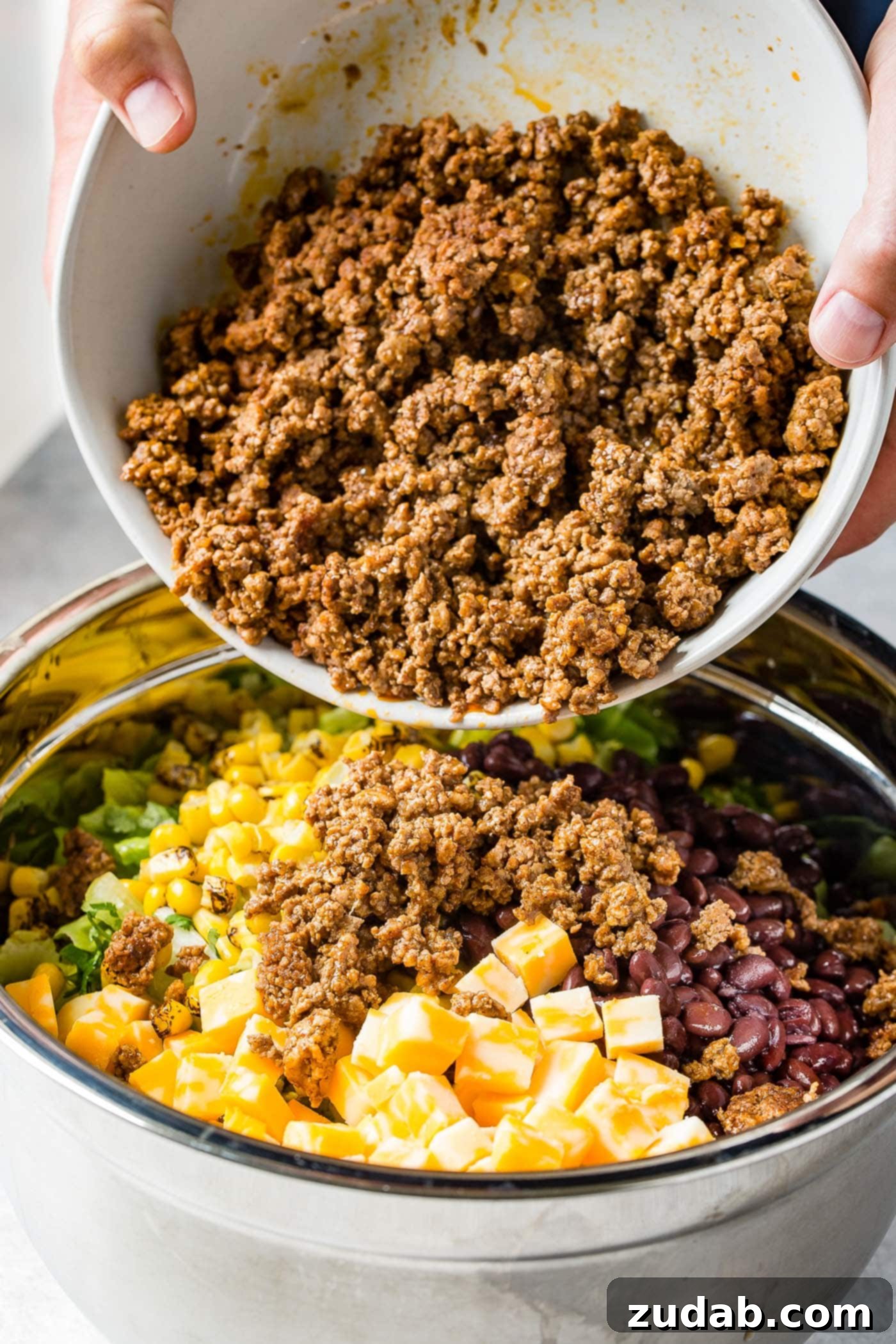 Seasoned taco meat being carefully poured into a large bowl, mingling with roasted corn, cheese cubes, tangy tomatillos, and freshly chopped onions, forming the savory base of a Tex-Mex taco salad.