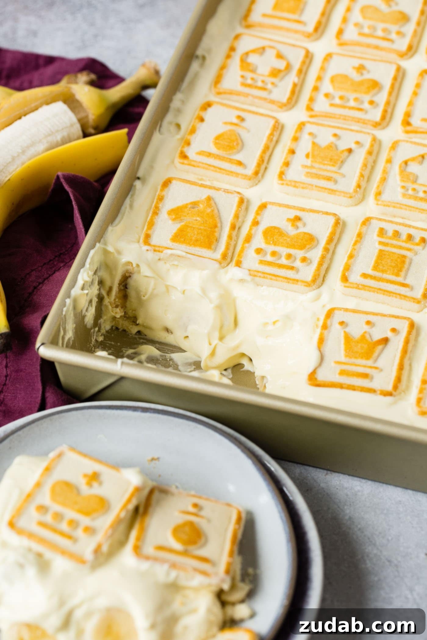 Paula Deen Banana Pudding A baking dish filled with Paula Deen's Not Yo Momma's Banana Pudding, showcasing its inviting layers. A serving has been removed, revealing the creamy texture and banana slices beneath. The top is covered with golden-brown Chessman cookies, and a ripe peeled banana rests beside the dish, highlighting the fresh fruit component of this delightful dessert.