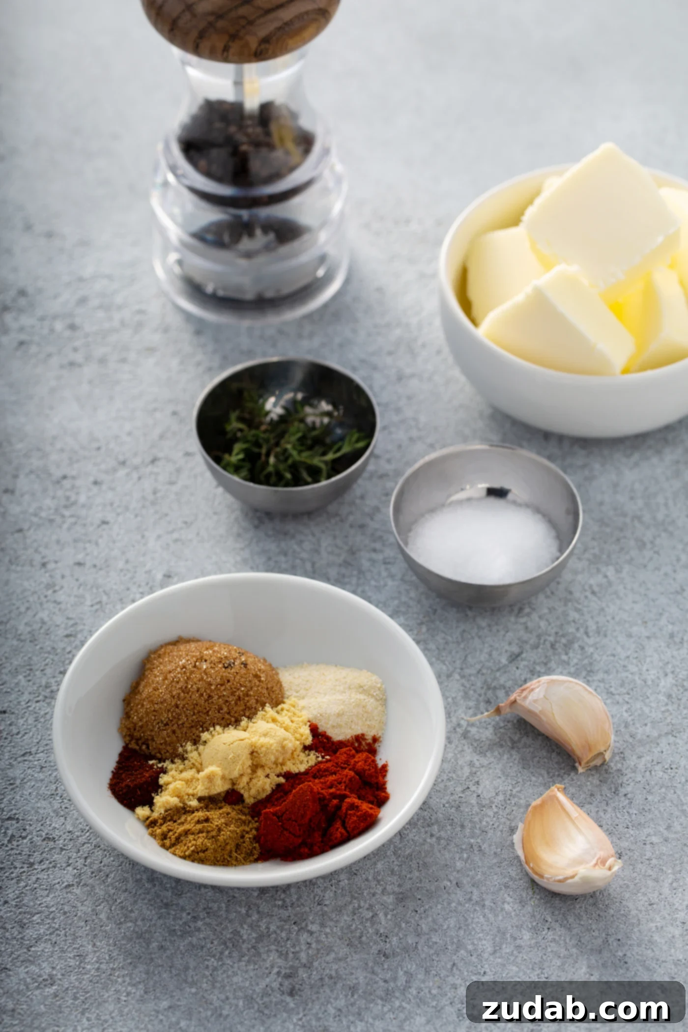 Small containers holding cubed butter, salt, and pepper are arranged on a wooden surface alongside a bowl containing distinct mounds of brown sugar, paprika, onion powder, cayenne pepper, and ground mustard. Two cloves of garlic complete the ingredient display.