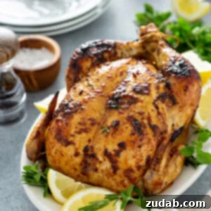 A whole roasted chicken on a white platter with lemon wedges and fresh thyme. On the table next to the chicken is a pepper shaker, salt in a wooden bowl, a stack of 3 dinner plates, and fresh thyme.