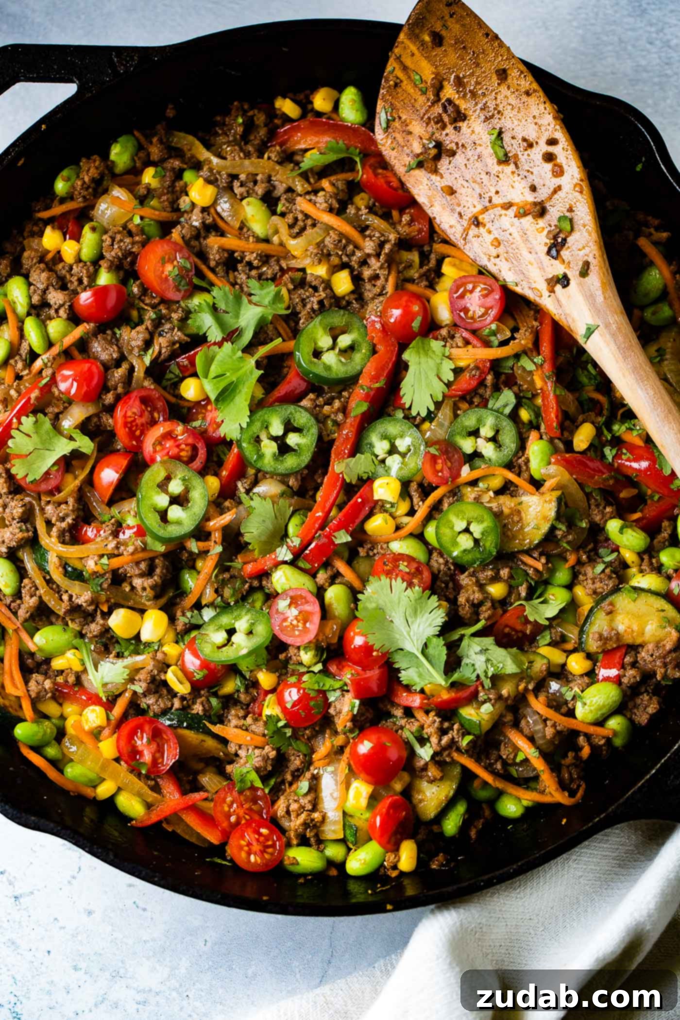 Fiesta Skillet Stir-Fry 3 A cast iron skillet filled with a colorful taco stir-fry, featuring ground beef, pasta, sliced tomatoes, jalapenos, carrots, and corn, with a wooden spoon resting on top.