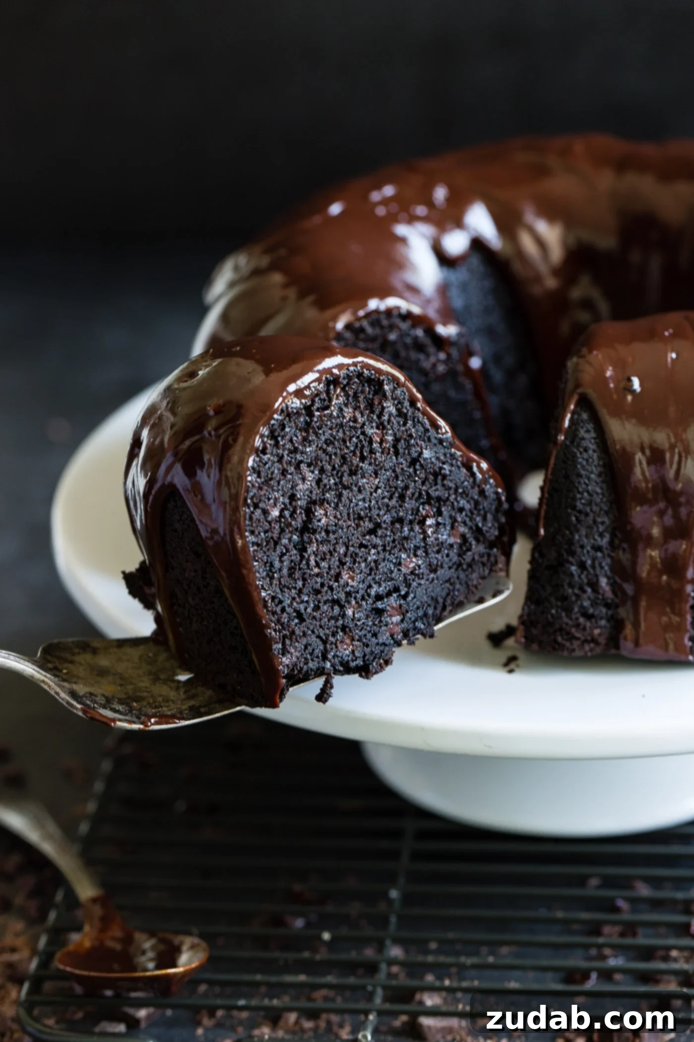 Moist Zucchini Dark Chocolate Bundt Cake A slice of zucchini chocolate bundt cake being removed from the cake. The inside of the cake is dark, moist chocolate and there is a chocolate glaze on top of the cake.