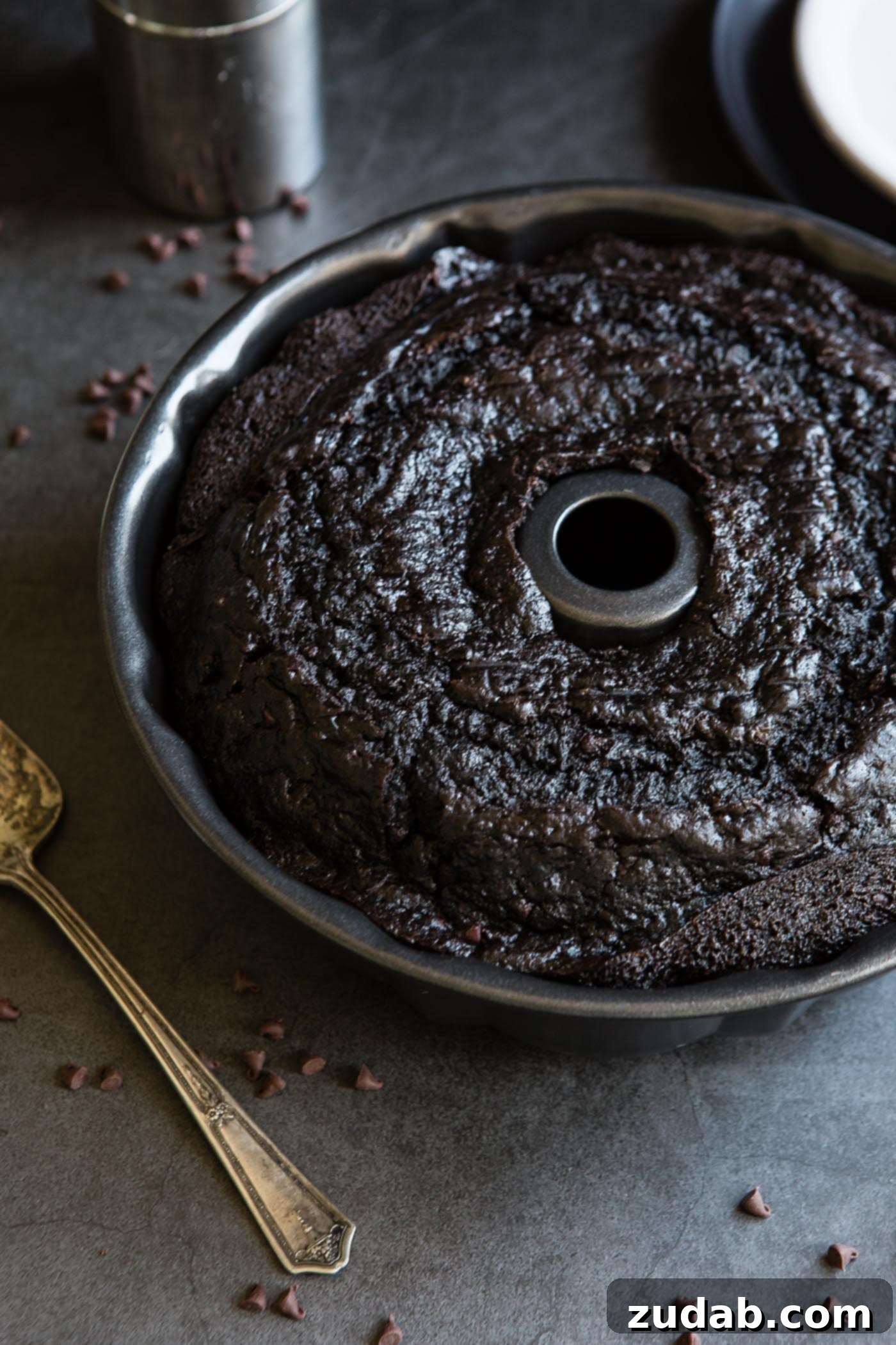 Moist Zucchini Dark Chocolate Bundt Cake A baked zucchini chocolate bundt cake in the bundt pan. Bits of chocolate and a cake server are next to the cake.