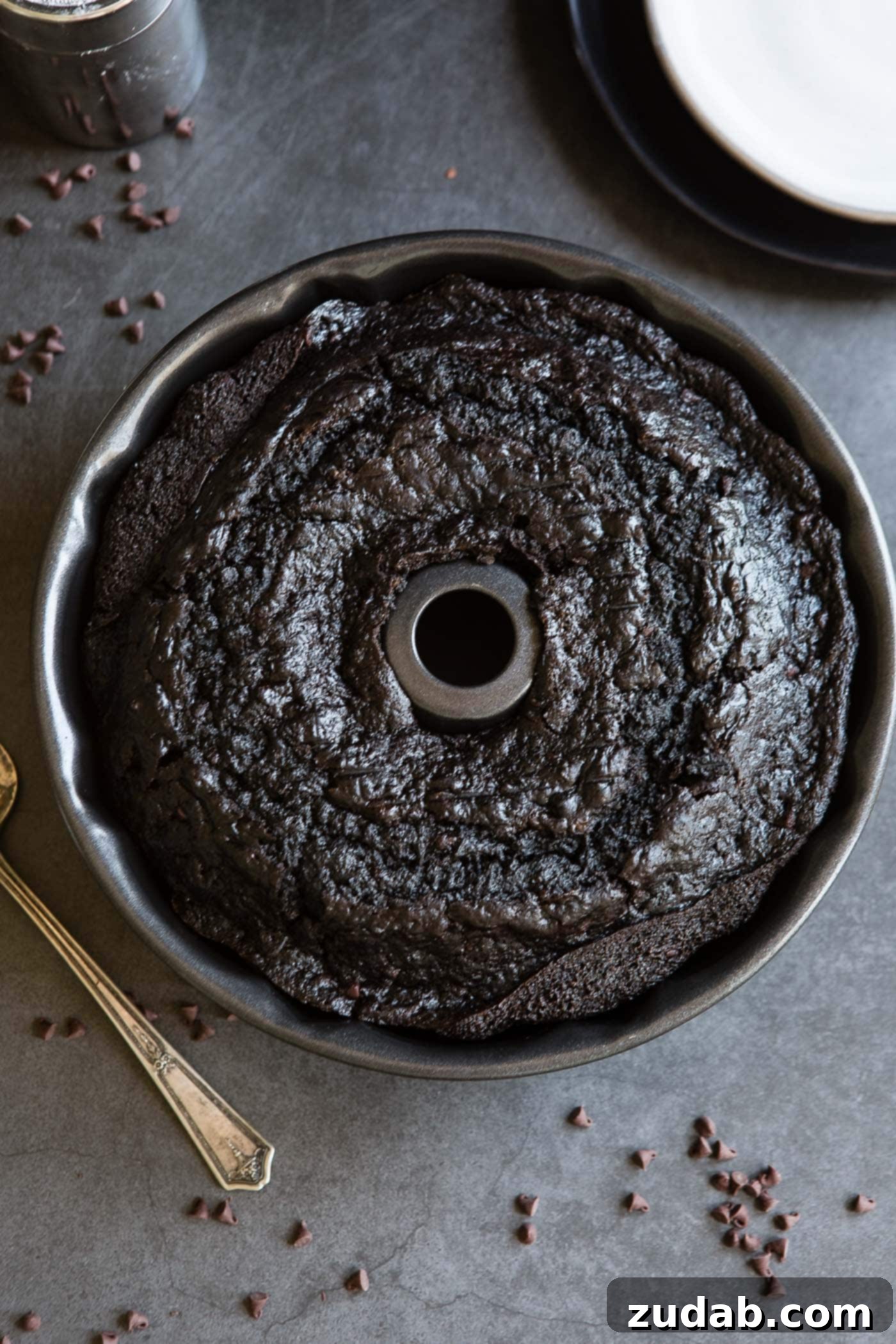 Moist Zucchini Dark Chocolate Bundt Cake A baked zucchini chocolate bundt cake in a bundt pan. A cake server and mini chocolate chips are on the table next to the cake.