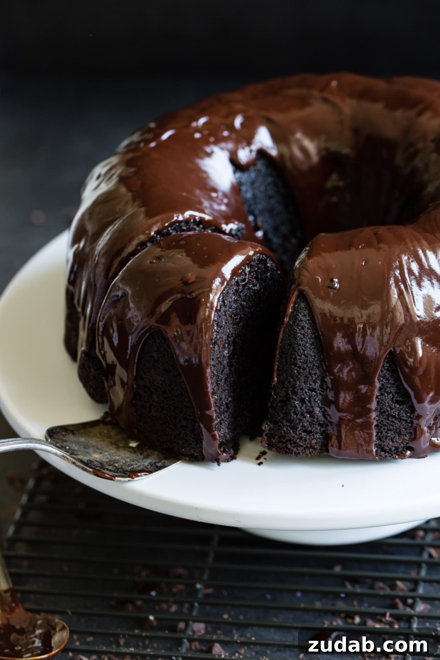 Moist Zucchini Dark Chocolate Bundt Cake A slice of cocoa veggie cake being removed from the whole bundt cake. The cake has a dark glaze on it.