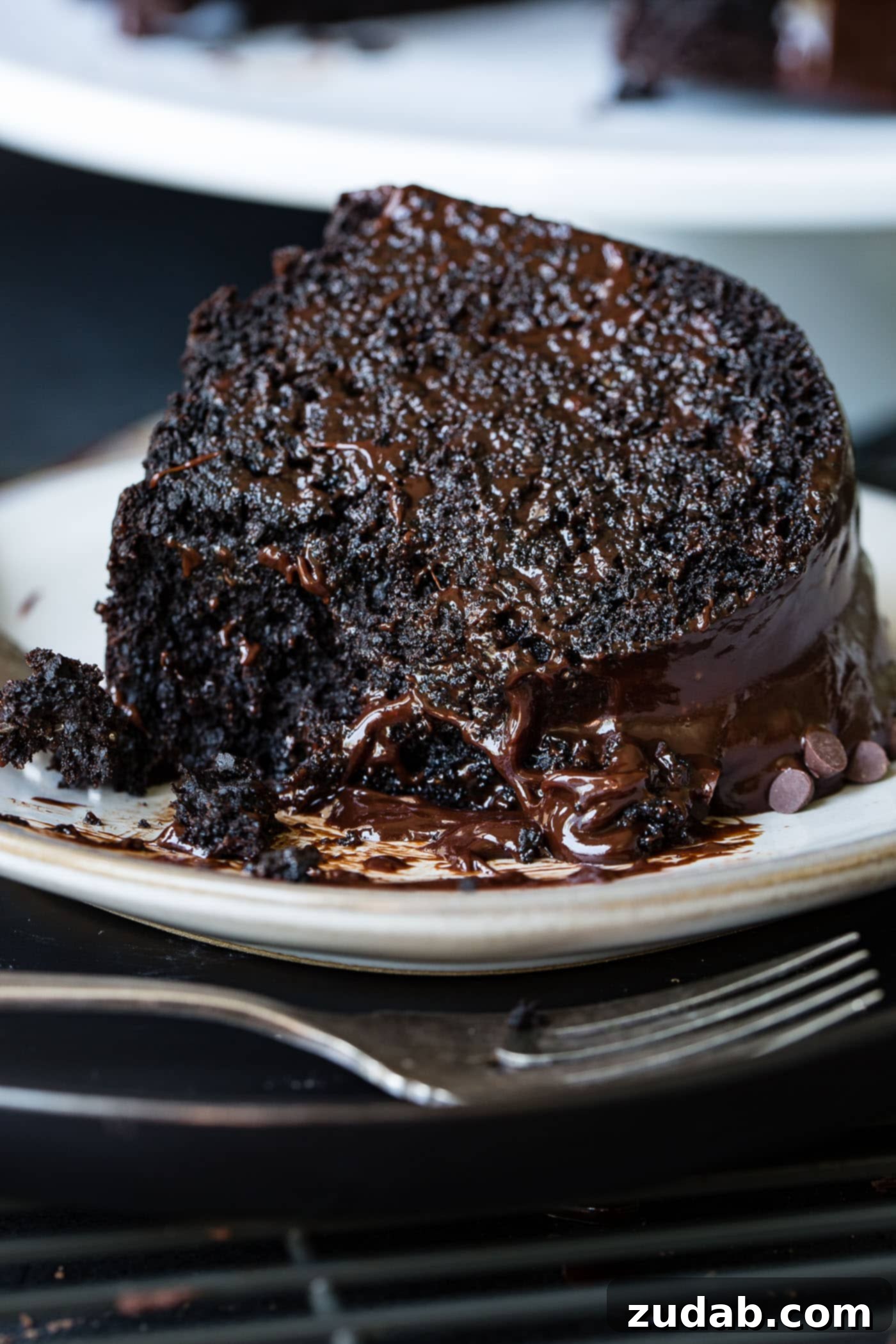 Moist Zucchini Dark Chocolate Bundt Cake A slice of zucchini chocolate bundt cake on a white dessert plate. The cake is dark, moist chocolate with a chocolate glaze. A fork is next to the plate.