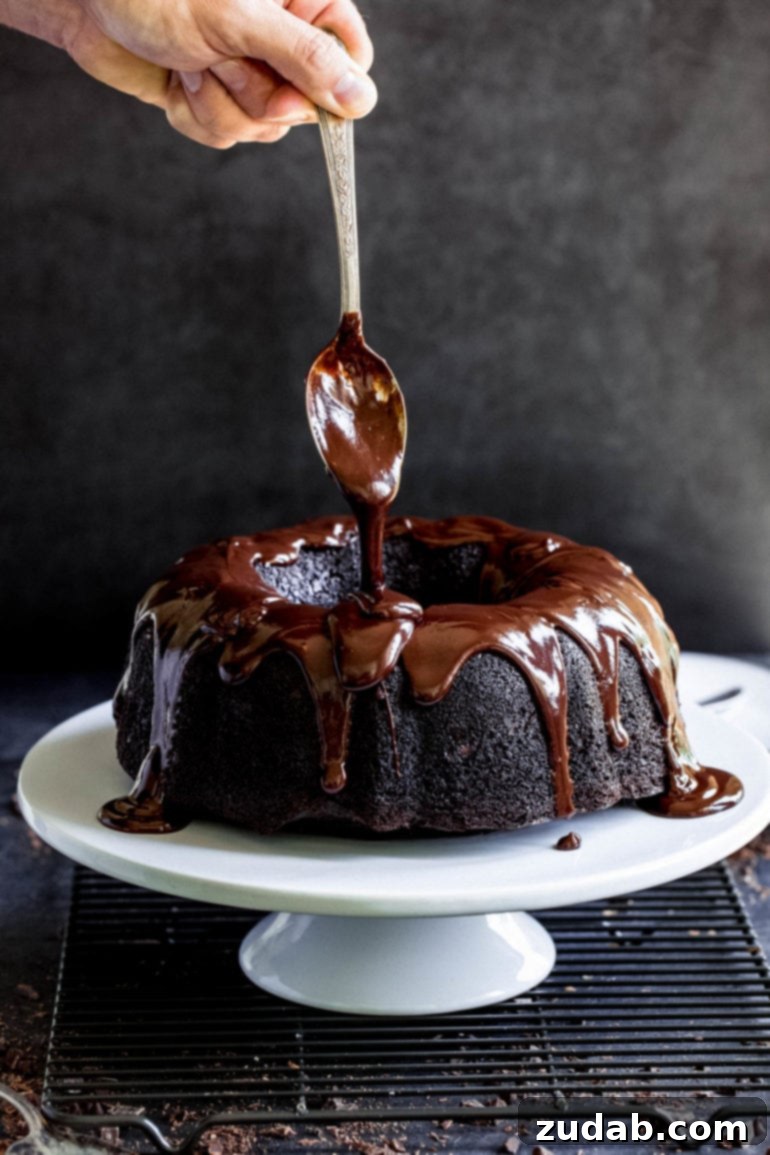 Fudgy Dark Chocolate Zucchini Bundt 2 A spoon with thick cocoa glaze drizzling over the top of a cake which is on a white cake plate.