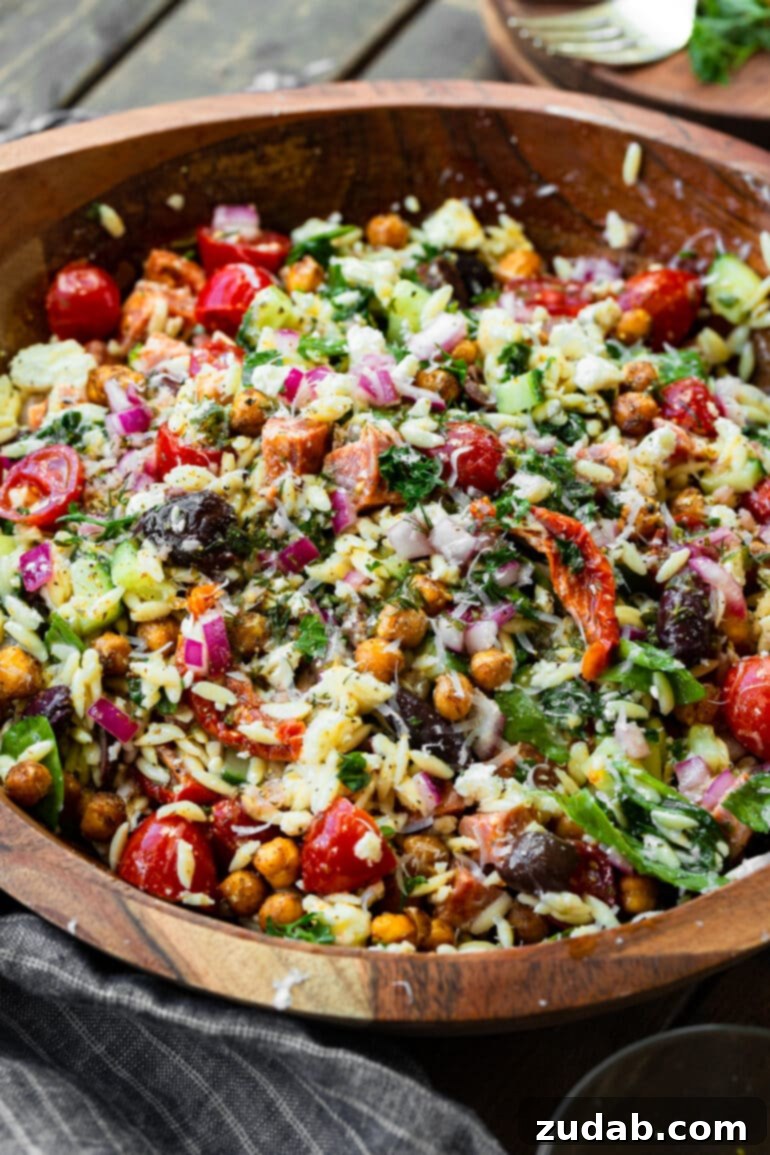 Zesty Mediterranean Orzo Salad 4 A close-up photo of a large wooden serving bowl filled with a colorful Greek orzo pasta salad, showcasing roasted chickpeas, sun-dried tomatoes, red onions, olives, and fresh herbs.