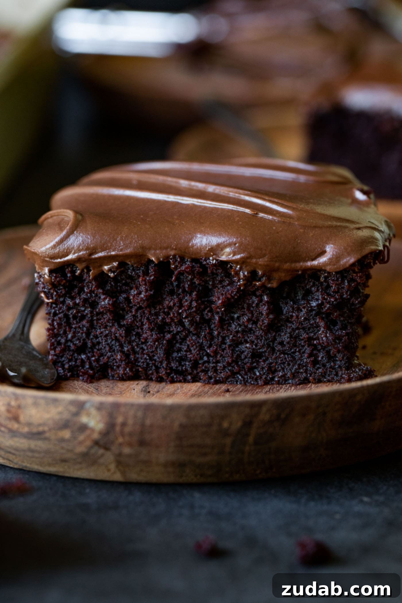 Moist Chocolate Zucchini Cake A slice of moist chocolate zucchini cake on a rustic wooden dessert plate. The cake is generously frosted with creamy chocolate cream cheese frosting, showing its rich texture.
