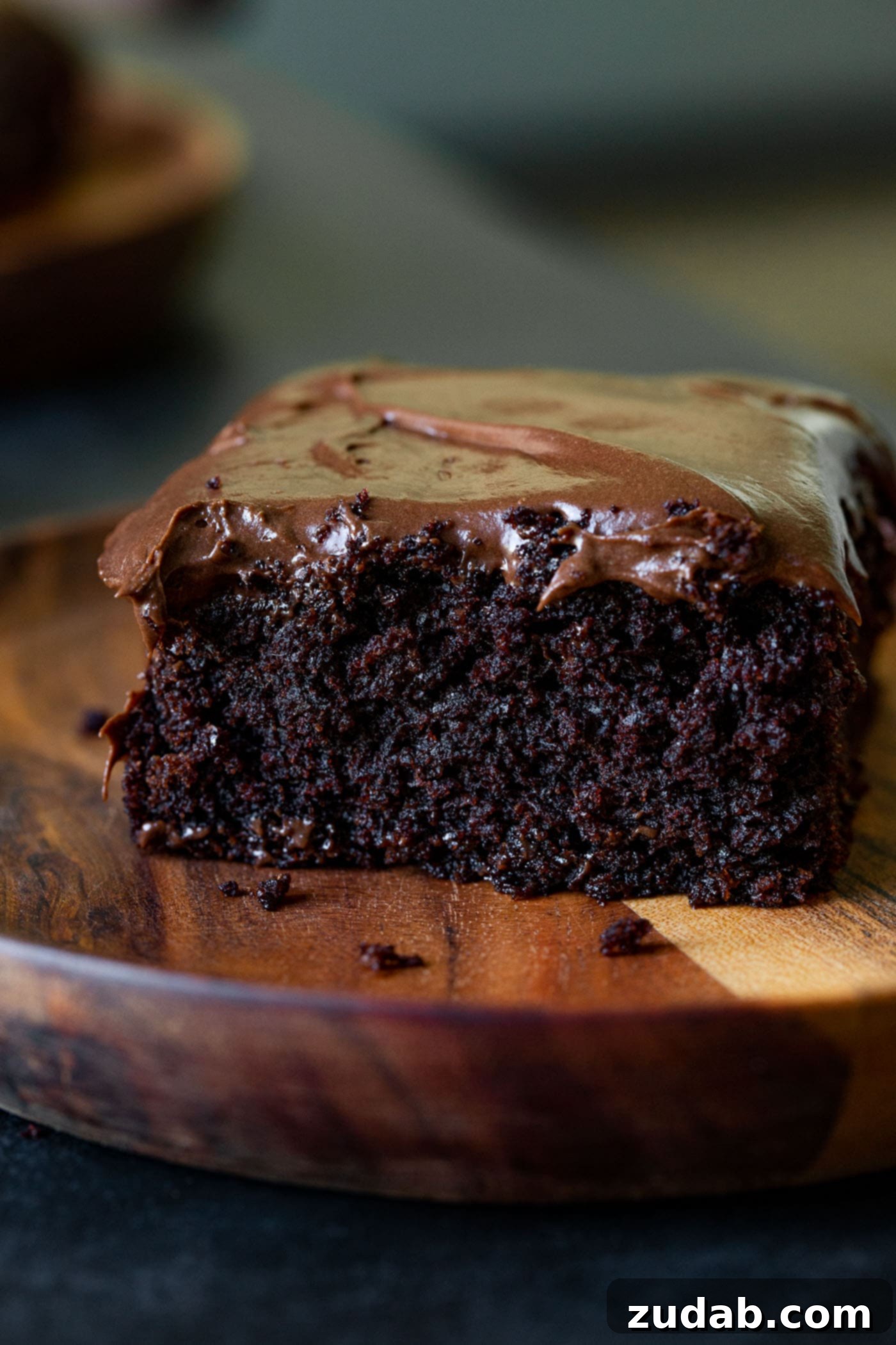 Moist Chocolate Zucchini Cake A close-up slice of moist chocolate zucchini cake, generously frosted with creamy chocolate cream cheese frosting, presented on a wooden dessert plate. The focus is on the rich, decadent texture.