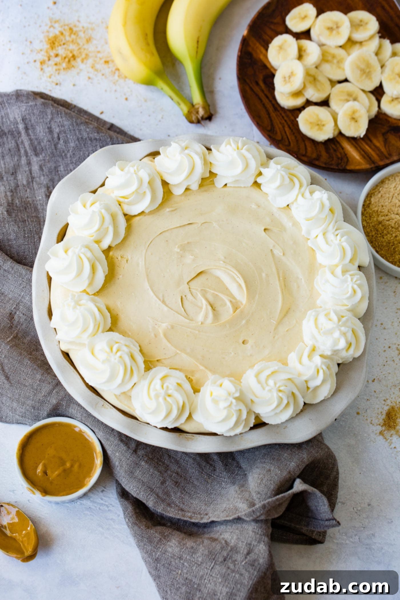 Peanut Butter Banana Cream Pie A peanut butter banana cream pie. Whipped cream has been piped around the edge of the pie and a cup of peanut butter, slices of banana, and a couple of bananas are in the background.