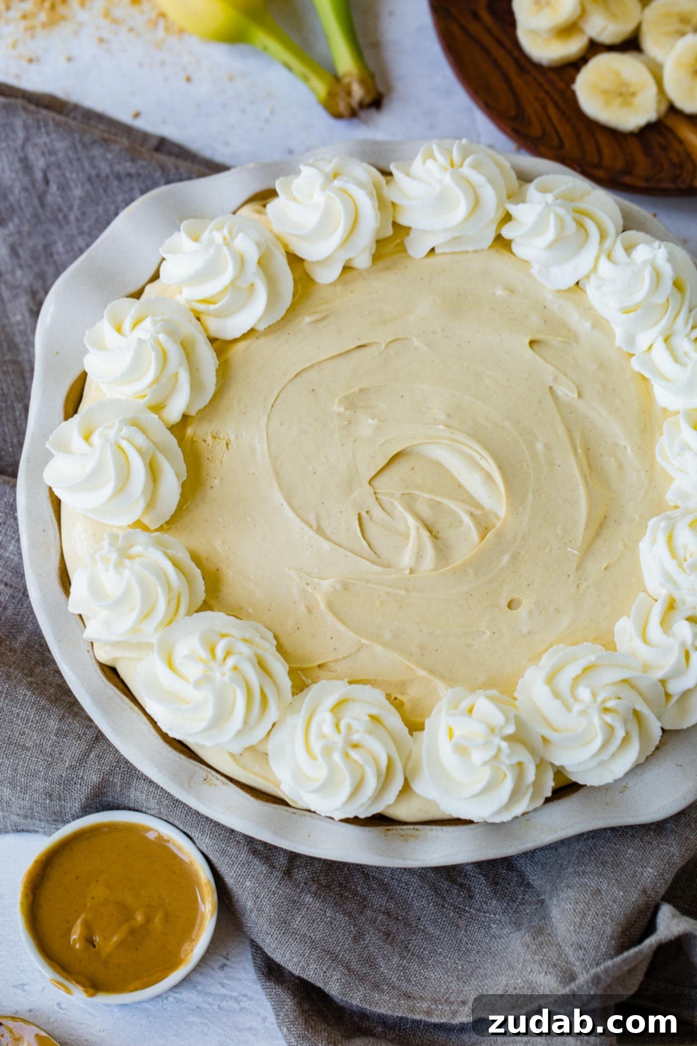 Peanut Butter Banana Cream Pie A peanut butter banana cream pie edged with whipped cream. A coup of peanut butter is next to the pie, and slices of banana are in the background.
