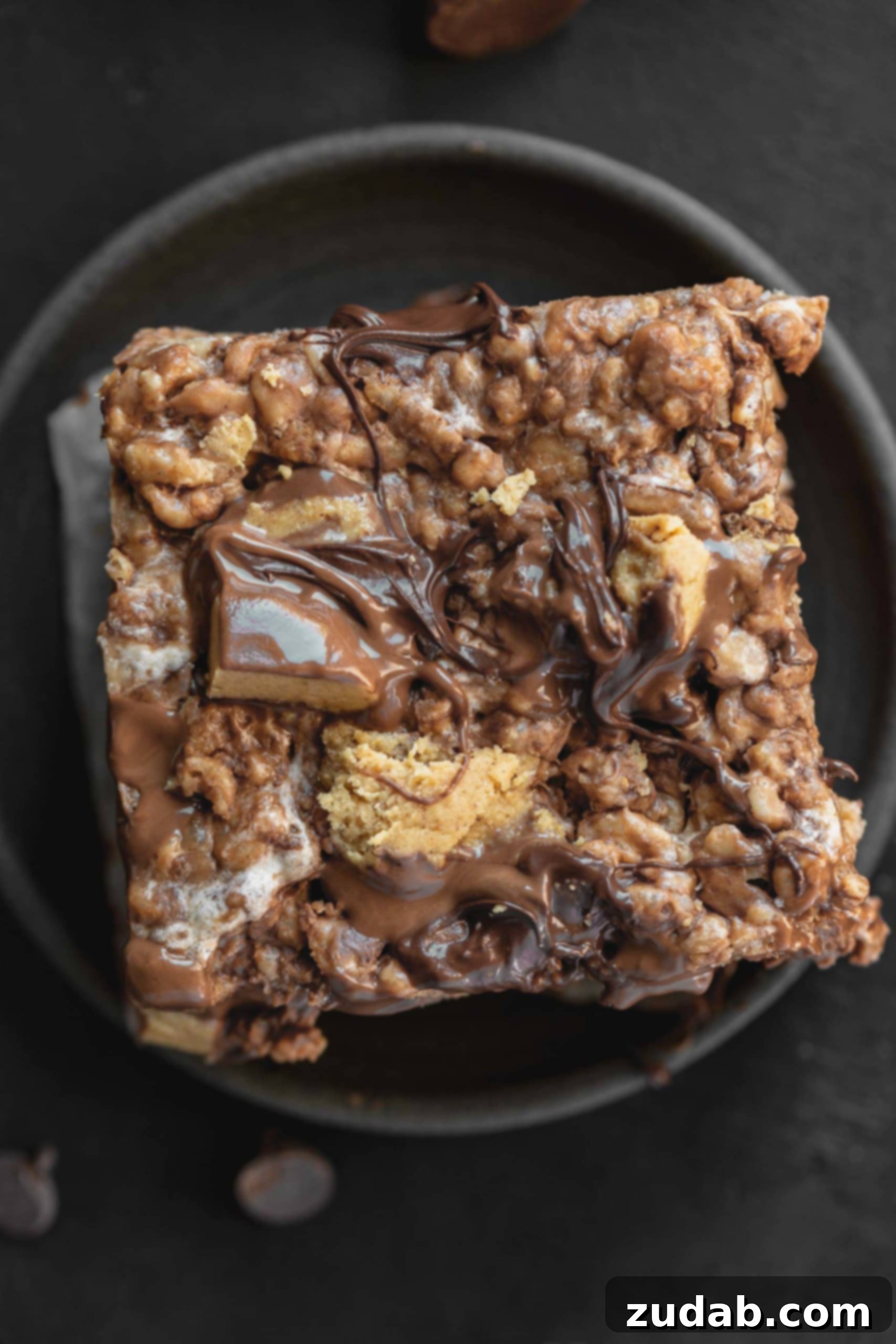 Reese's Rice Krispies Treats A photo of a single peanut butter rice krispies treat topped with chunks of chopped Reese's Peanut Butter Cups and melted chocolate chips on a dark gray plate.