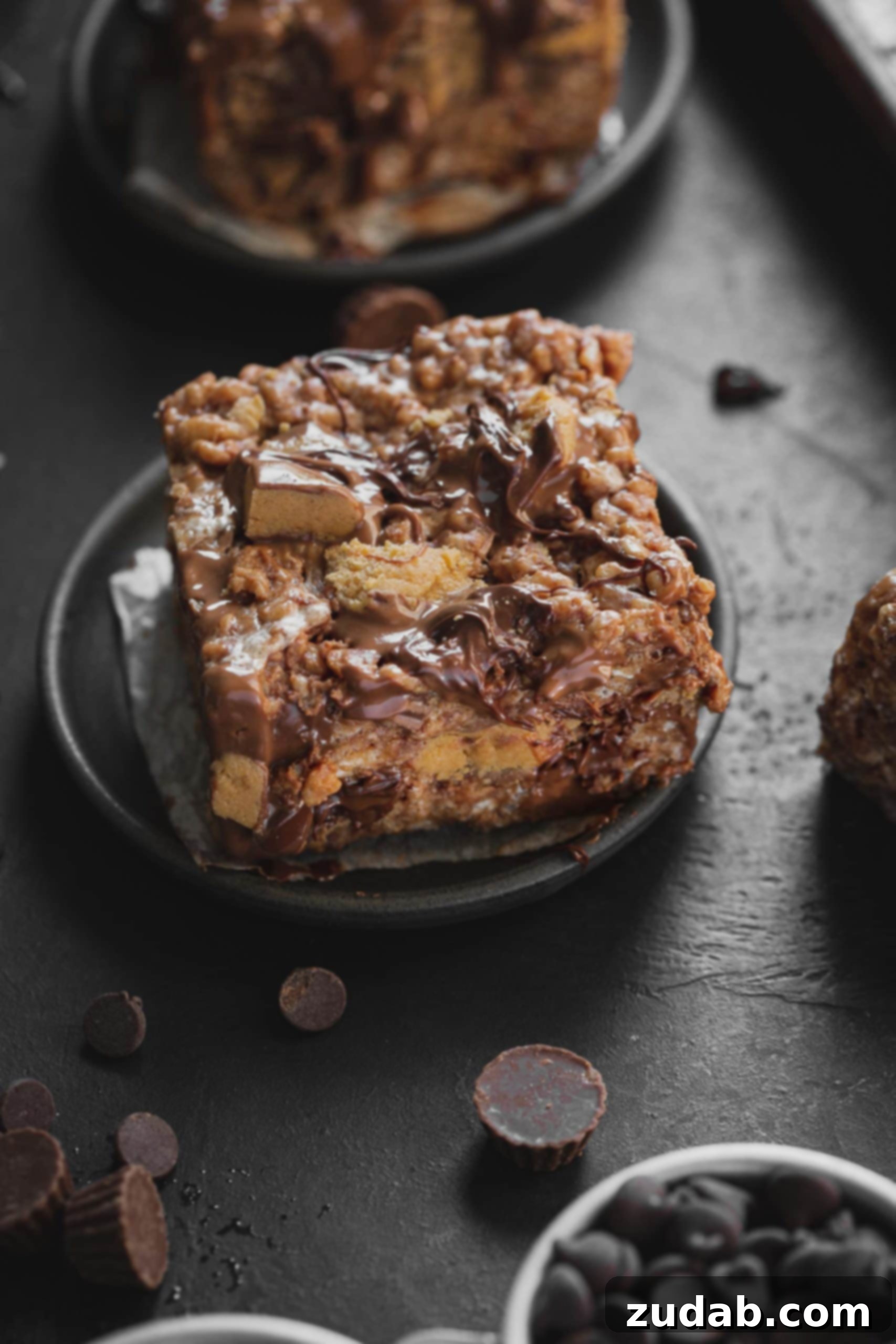 Reese's Rice Krispies Treats A photo of a single peanut butter rice krispies treat loaded with chunks of Reese's Peanut Butter Cups and melted chocolate chips on a small dark gray plate, with another rice krispies treat in the background.
