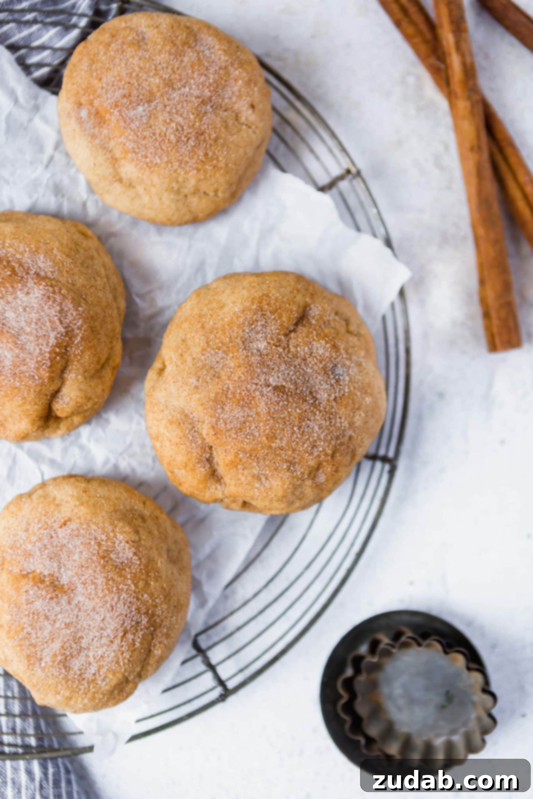Baking Thick Snickerdoodle Cookies at Home A cooling rack filled with golden-brown thick snickerdoodle cookies, each generously sprinkled with cinnamon sugar. A few fragrant cinnamon sticks rest beside the rack, adding to the inviting aroma.