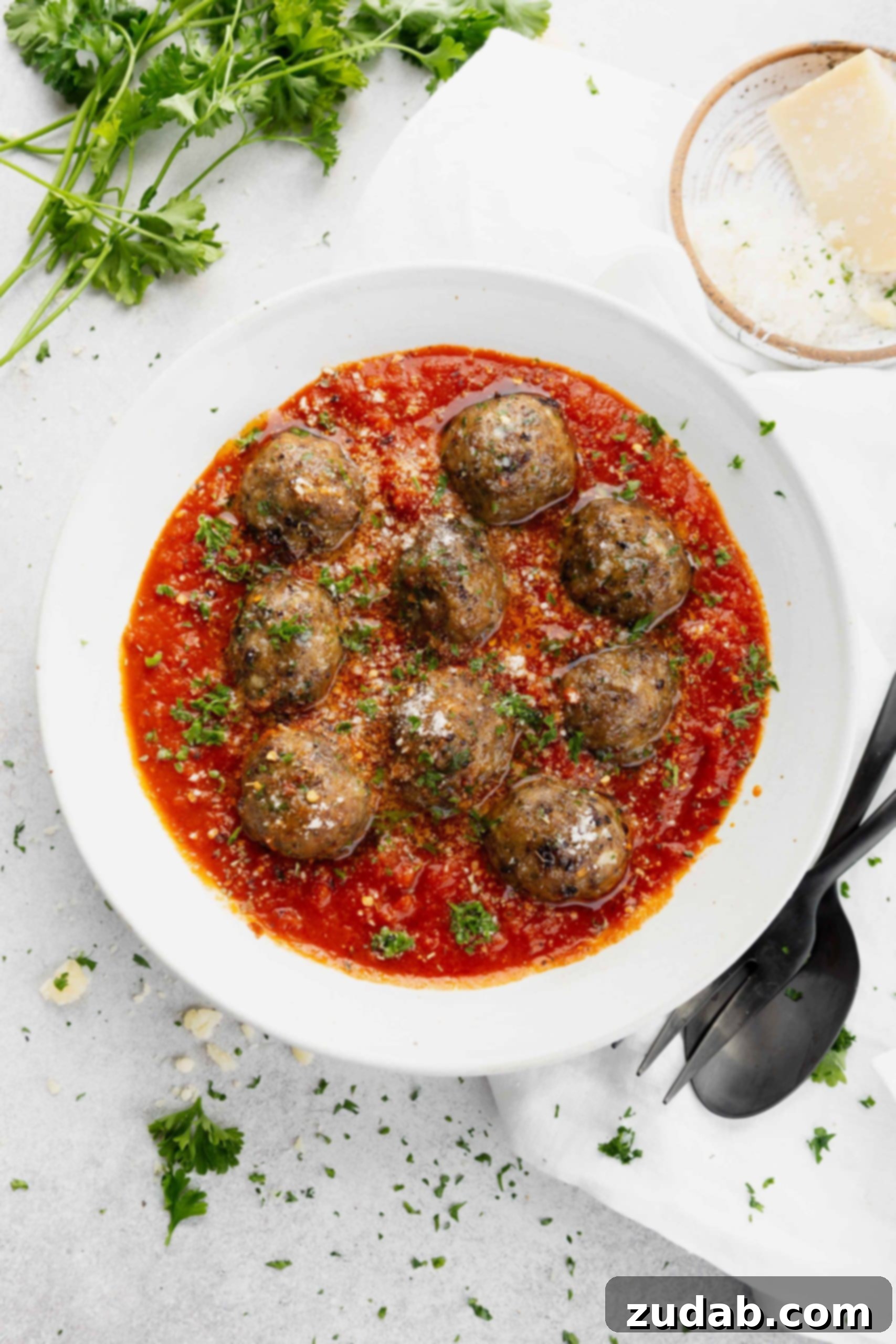 Melt in Your Mouth Meatball Recipe A bowl of tender meatballs coated in a rich tomato sauce, garnished with freshly chopped parsley. A fork and spoon rest beside the bowl, inviting a taste.