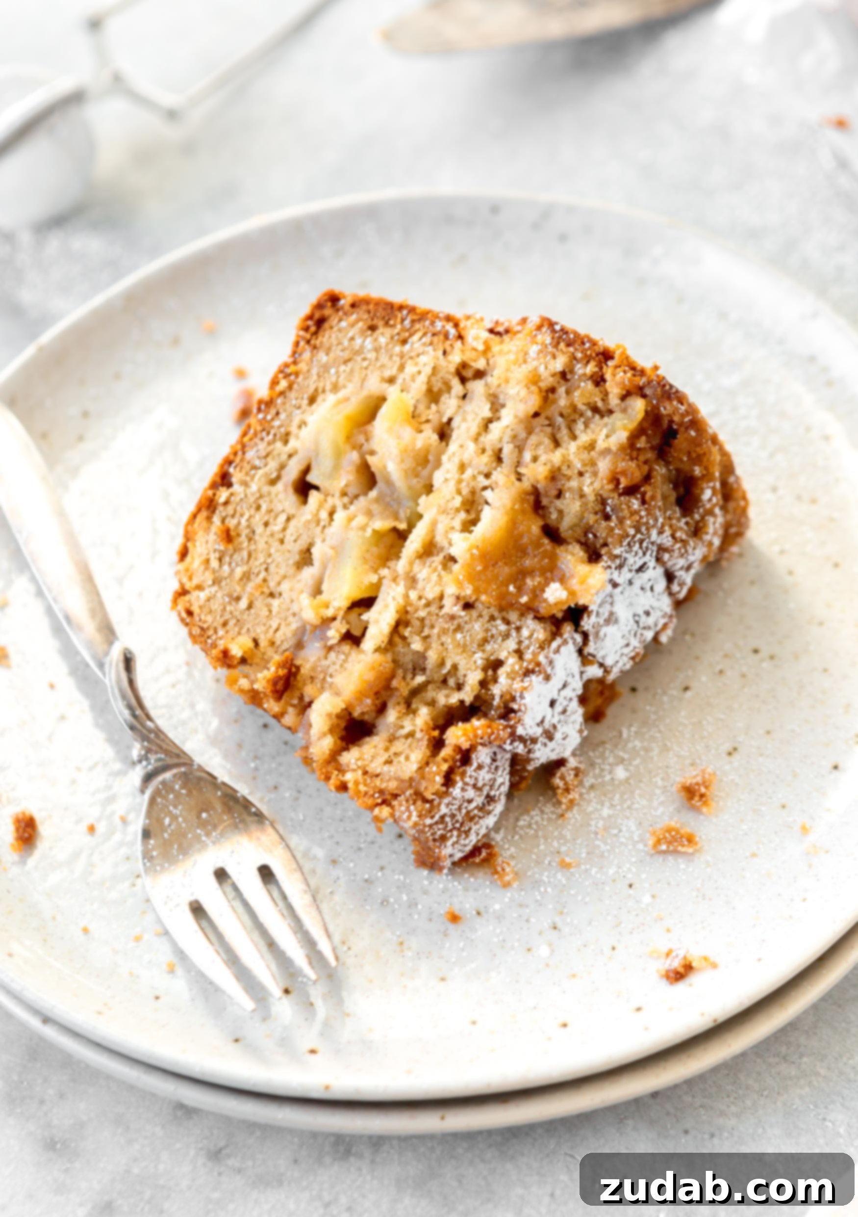 Jewish Apple Cake A slice of Jewish apple cake on a white dessert plate with a fork next to it. The cake is dense, and filled with apples. There is powdered sugar on the cake top.