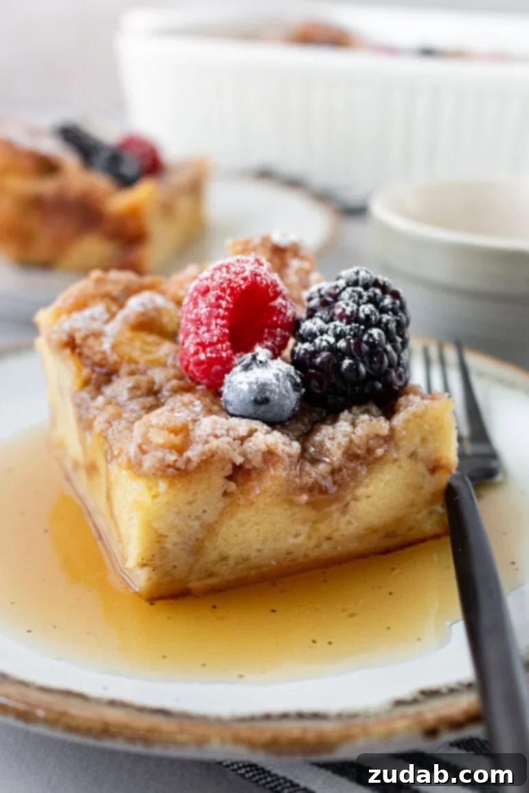 A slice of French toast casserole on a plate, drenched in maple syrup, with a fork poised for a bite and fresh berries scattered on top.