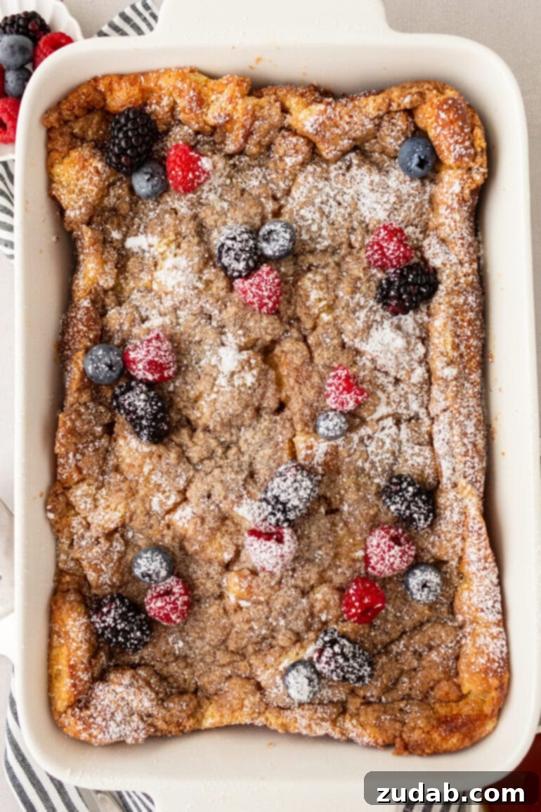 A golden-brown French toast casserole, adorned with a sweet streusel topping, powdered sugar, and a scattering of fresh berries, in a full baking dish.