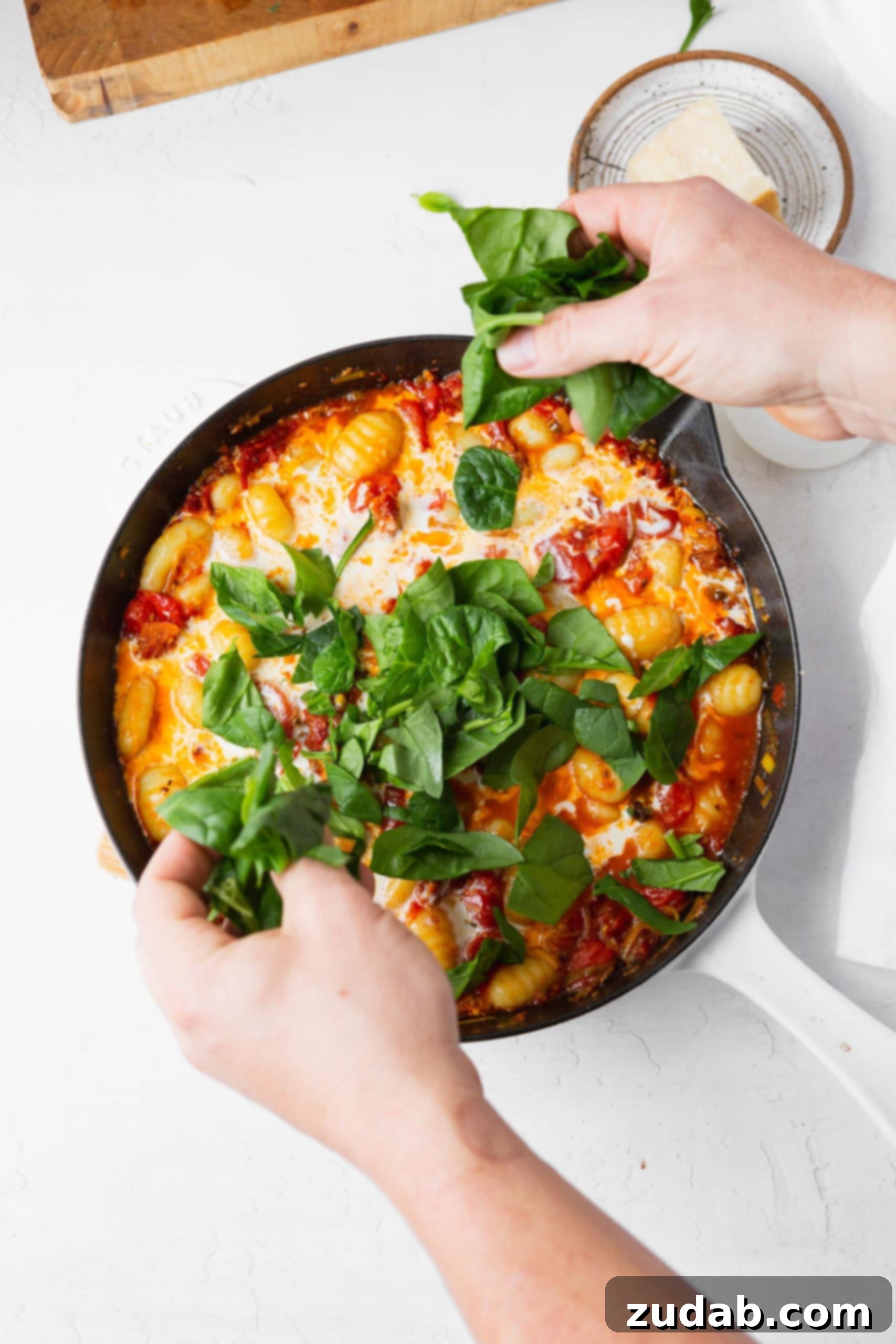 Adding Fresh Spinach to Gnocchi A hand sprinkling fresh spinach leaves into a cast iron skillet filled with creamy sun-dried tomato and spinach gnocchi, adding a vibrant touch.