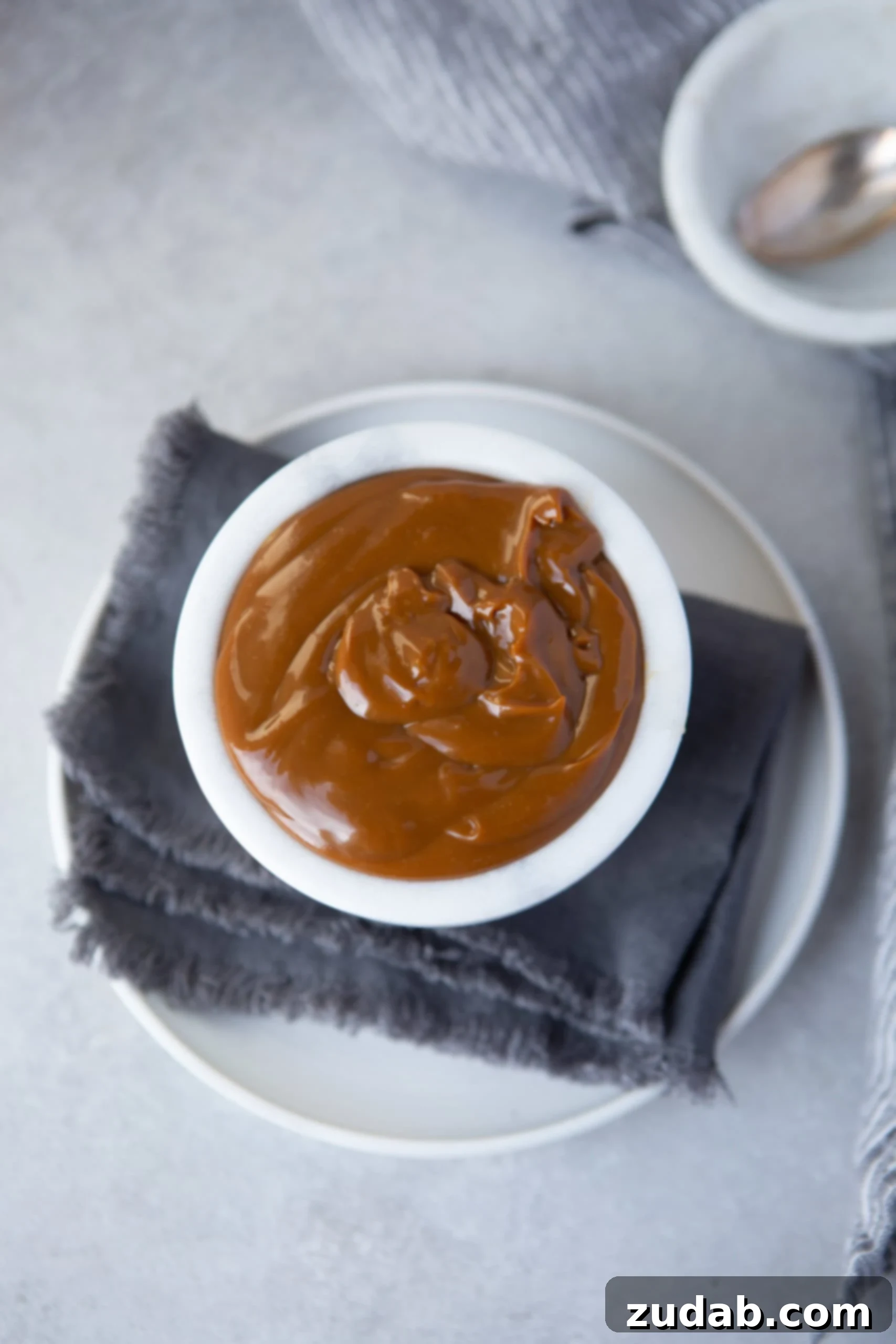 Homemade Dulce de Leche Recipe An overhead shot of a white cup filled with luxurious dulce de leche, set on a vibrant blue cloth napkin atop a pristine white plate.