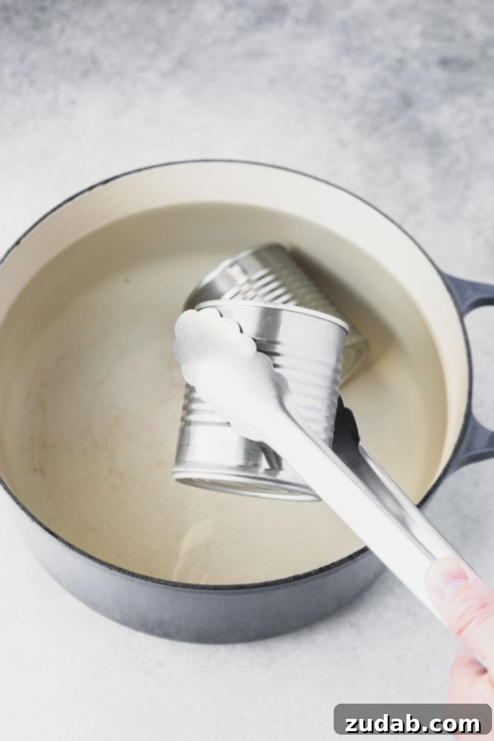 Homemade Dulce De Leche Recipe A pair of metal tongs carefully removing a sealed can of sweetened condensed milk from a pot of simmering water.