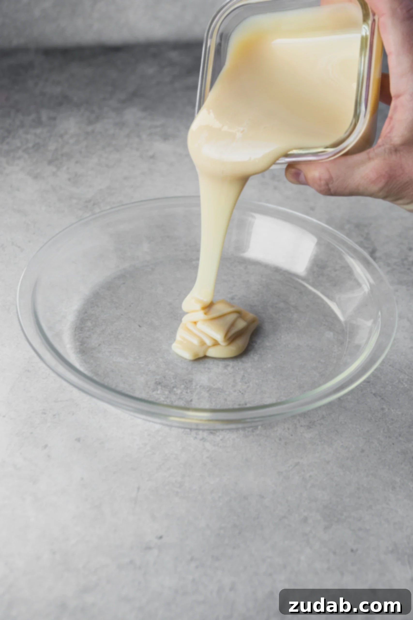 Homemade Dulce De Leche Recipe Sweetened condensed milk being poured from a can into a clear glass pie dish, ready for the oven method.