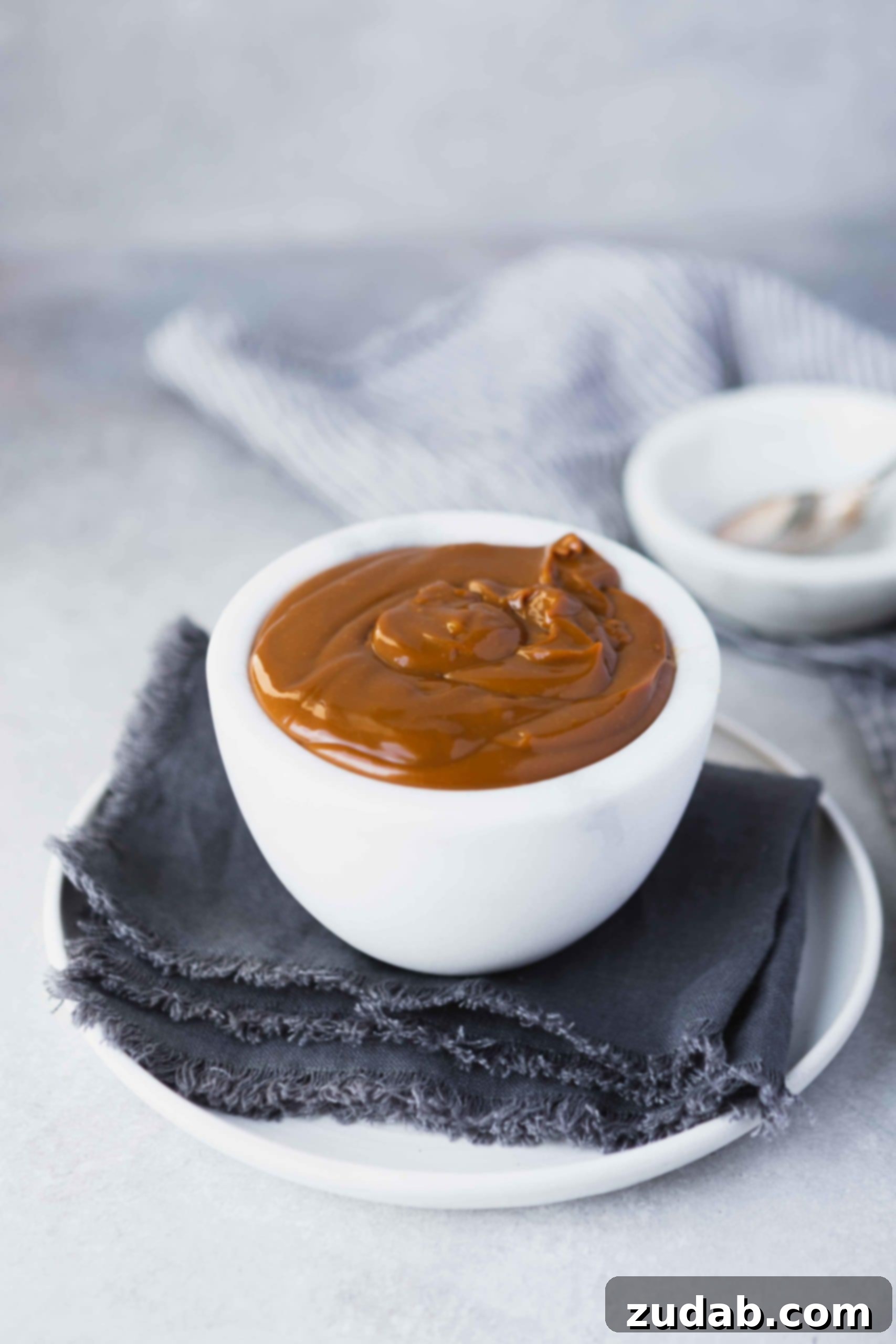 Homemade Dulce De Leche Recipe A pristine white cup filled with golden-brown dulce de leche, presented on a blue cloth napkin and a white plate, inviting a taste.