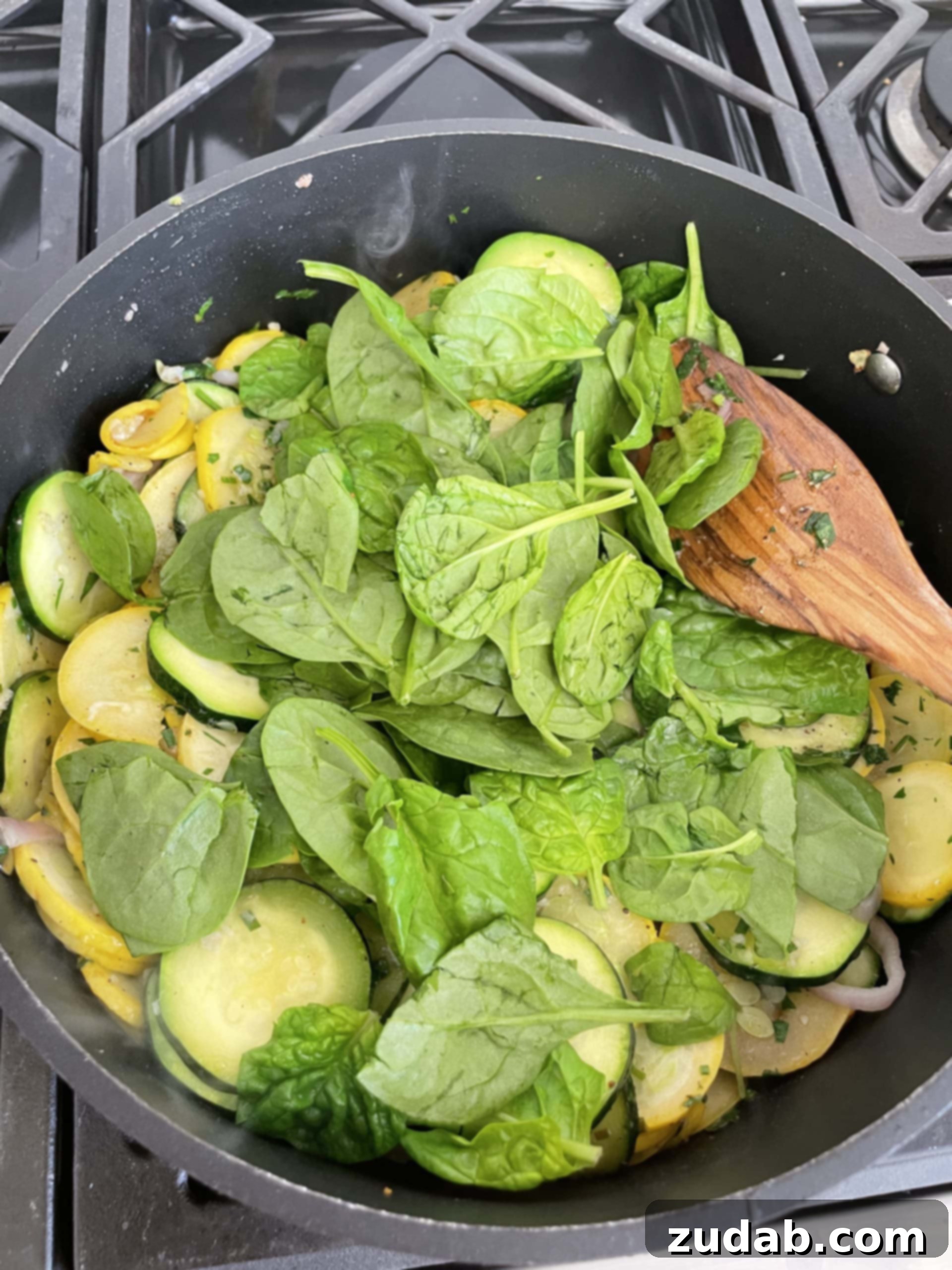 A cast iron skillet on the stovetop, with zucchini, yellow squash, and fresh spinach being stirred together with a wooden spoon, blending flavors and colors.