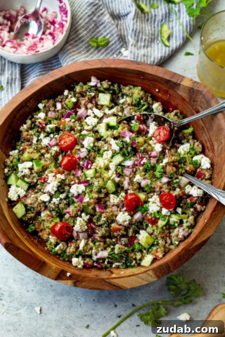 Mediterranean Quinoa Bowl 3 A large wooden serving bowl overflowing with Mediterranean Quinoa Salad, featuring a tantalizing mix of fresh vegetables, herbs, and feta, with two elegant silver serving spoons nestled within.