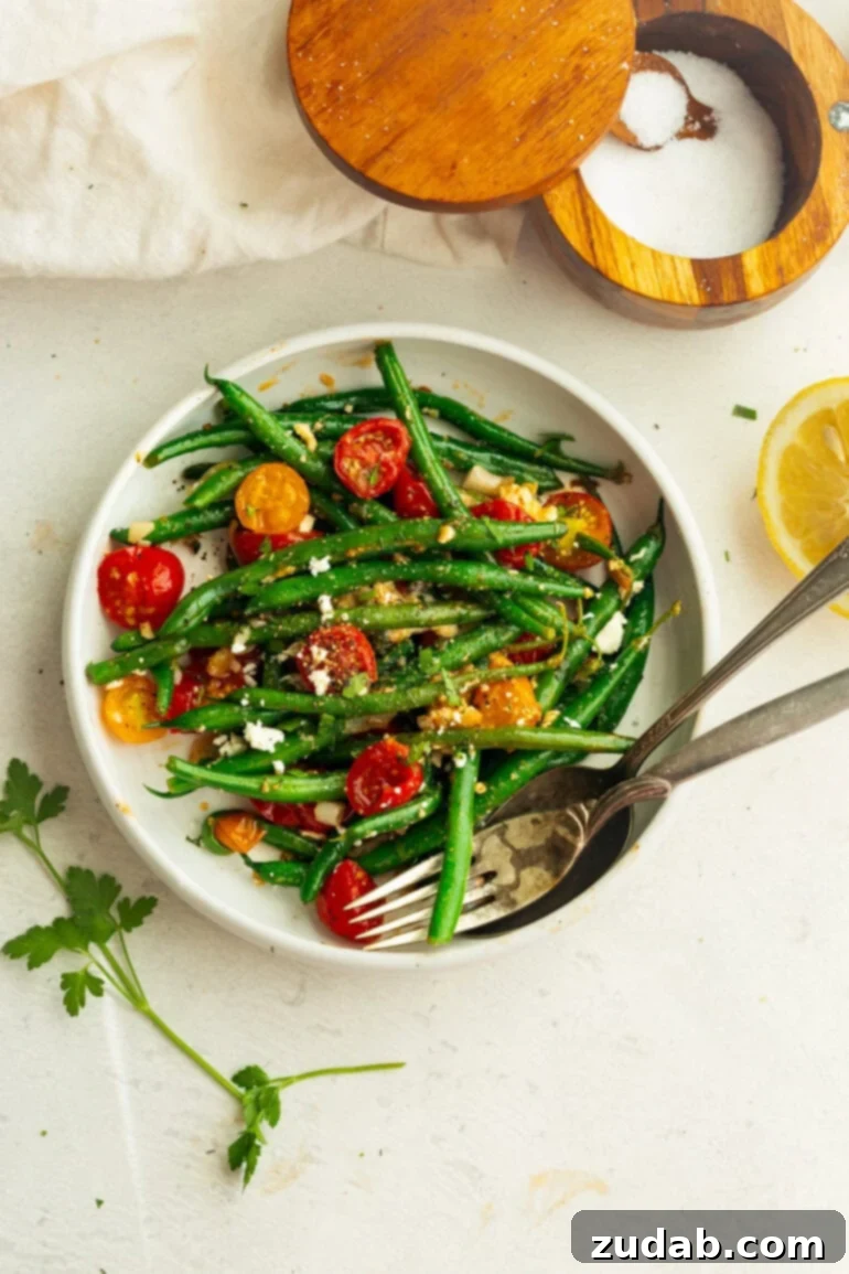 Garlic Green Bean and Cherry Tomato Sauté 4 A white bowl filled with glistening sautéed green beans and halved cherry tomatoes, topped with crumbled feta cheese and fresh herbs, with a fork resting inside.