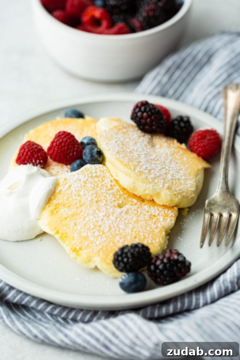 Pillowy Soft Japanese Pancakes 7 A photo of three Japanese cloud pancakes sitting on a plate topped with powdered sugar, mixed berries scattered around them, and a dollop of whipped cream on the side.