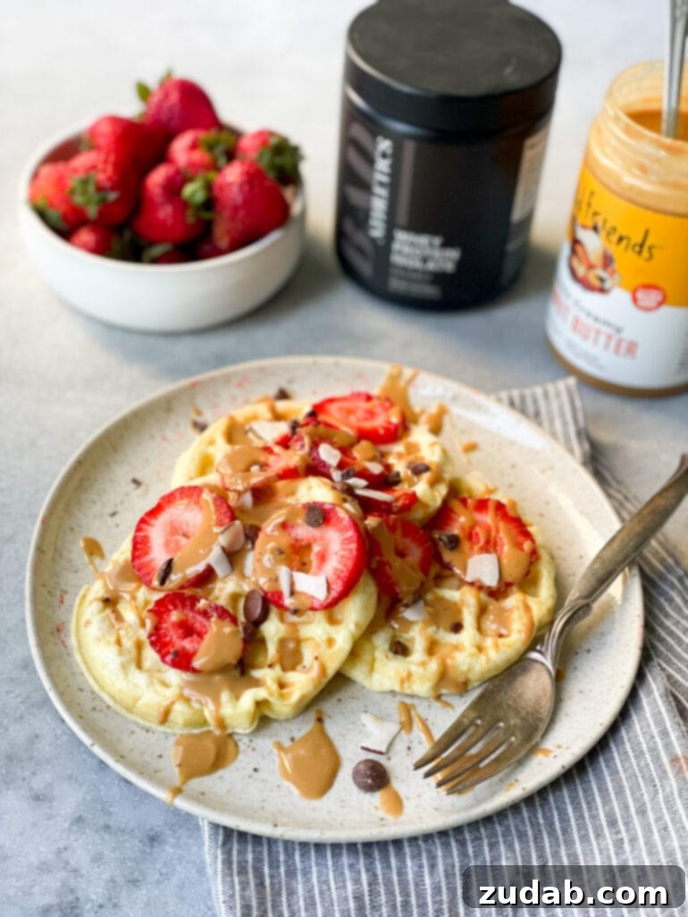 A beautiful photo featuring three small circular protein waffles on a white dinner plate, generously topped with sliced strawberries, chocolate chips, and a luscious peanut butter drizzle. In the background, a bowl of whole strawberries, a container of protein powder, and a jar of peanut butter complete the healthy breakfast scene.