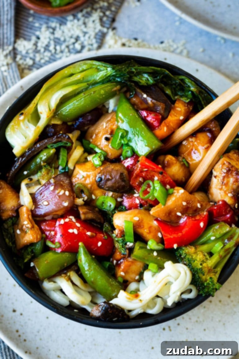 a close up photo of a bowl full of chicken stir fry with red bell peppers, broccoli, snap peas, mushrooms, bok choy and chunks of juicy chicken visible.